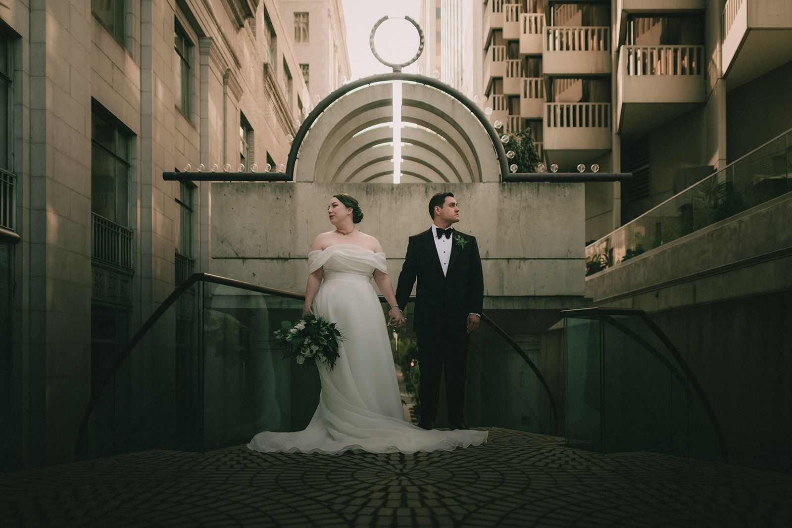 Anne and Charles stand hand in hand beneath geometric city architecture in San Francisco.