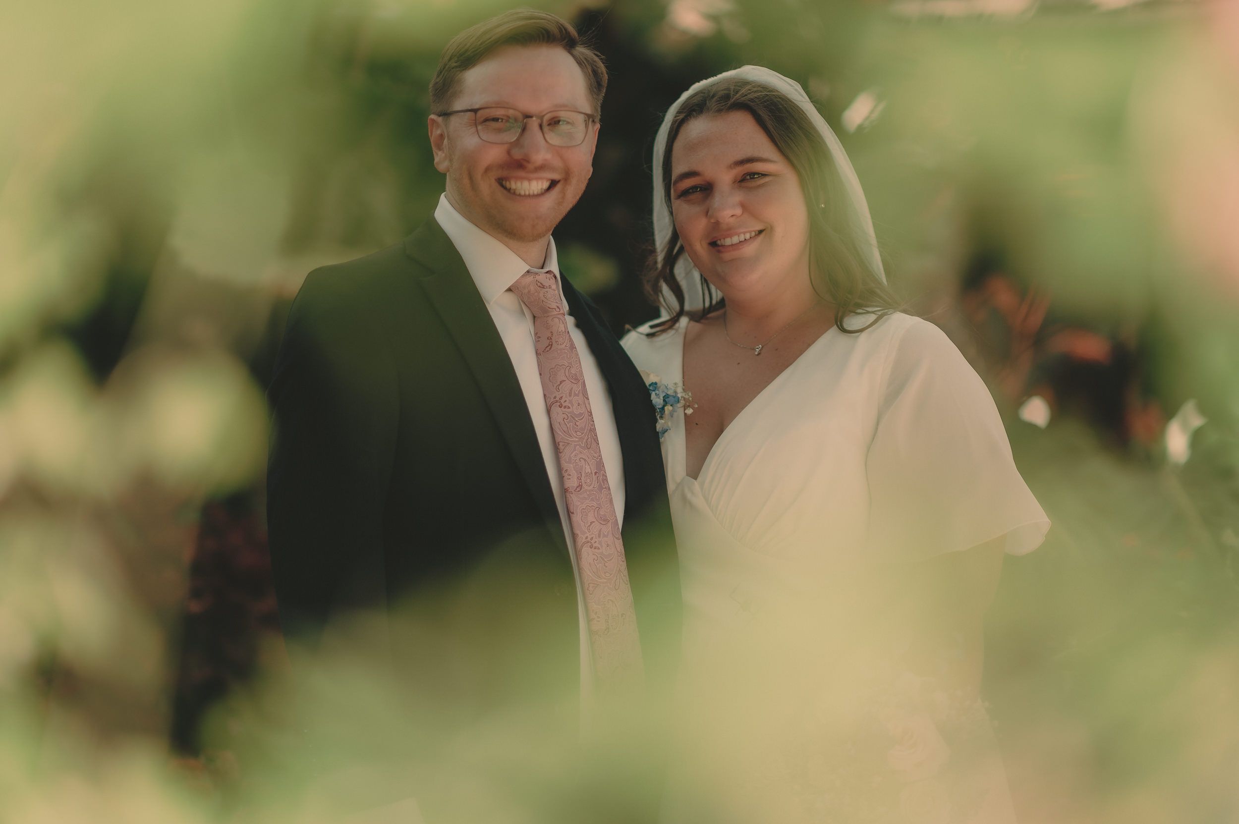Tiffani and Braden lean close behind soft green foreground leaves.
