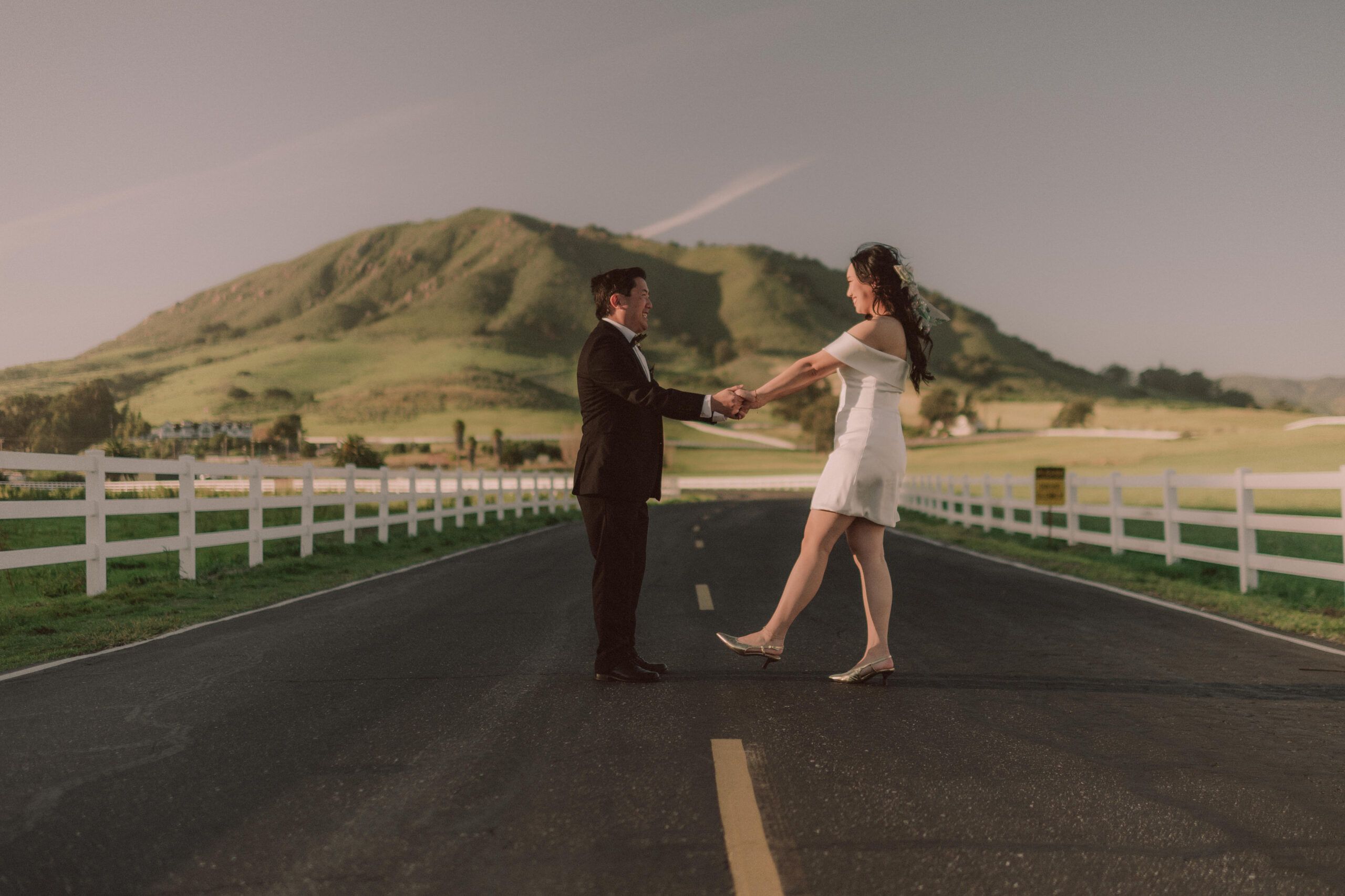 A newlywed couple dancing together on a country road with rolling hills behind them