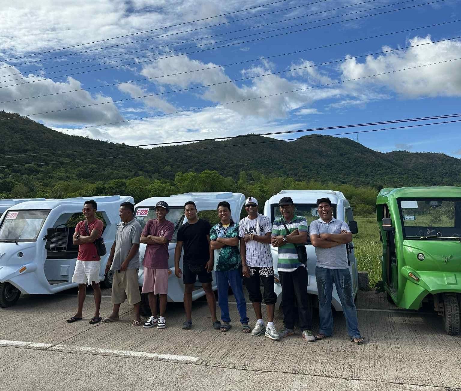 A group of seven smiling local public transport operators standing proudly in front of a fleet of white and green GerWeiss Motors GE4 electric tricycles. These drivers participate in the Boracay 100% EV transition program, utilizing a zero-cash-out battery swapping ecosystem that empowers the local community and increases daily take-home pay by 200 to 500 percent.