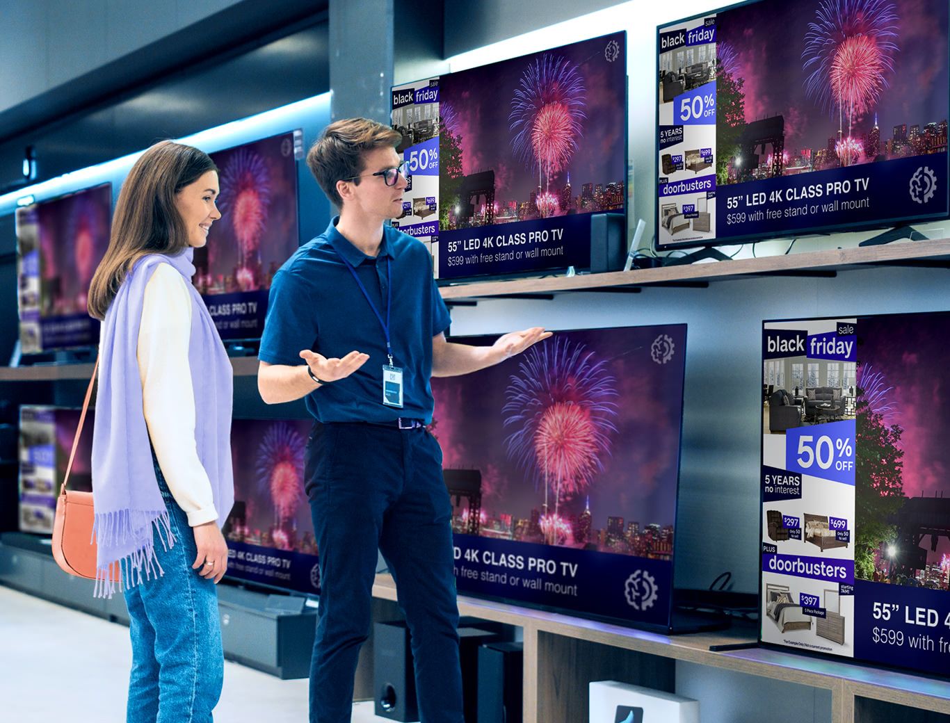 A woman speaking with a salesman while standing in front of a display wall of large TVs