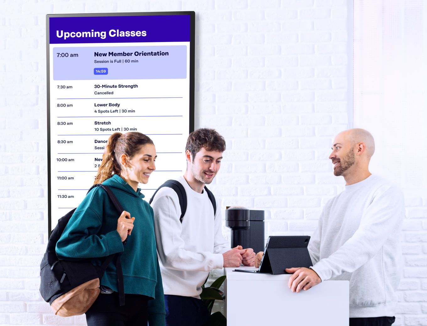 A woman and man at the front desk of a gym with a class schedule screen displayed behind them