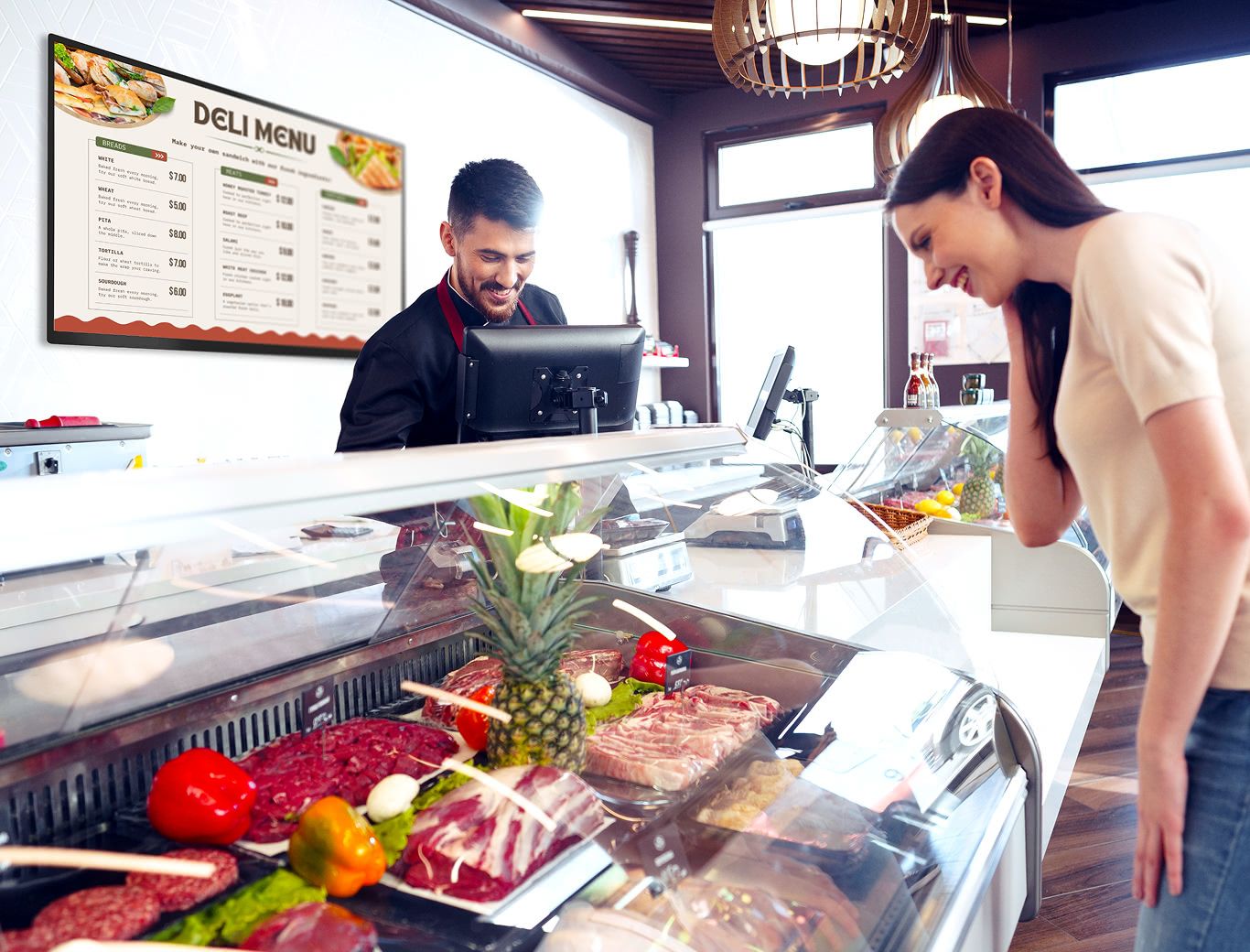 Woman at a deli counter looking over selections and chatting with the working behind the counter with a deli menu displayed in the background.