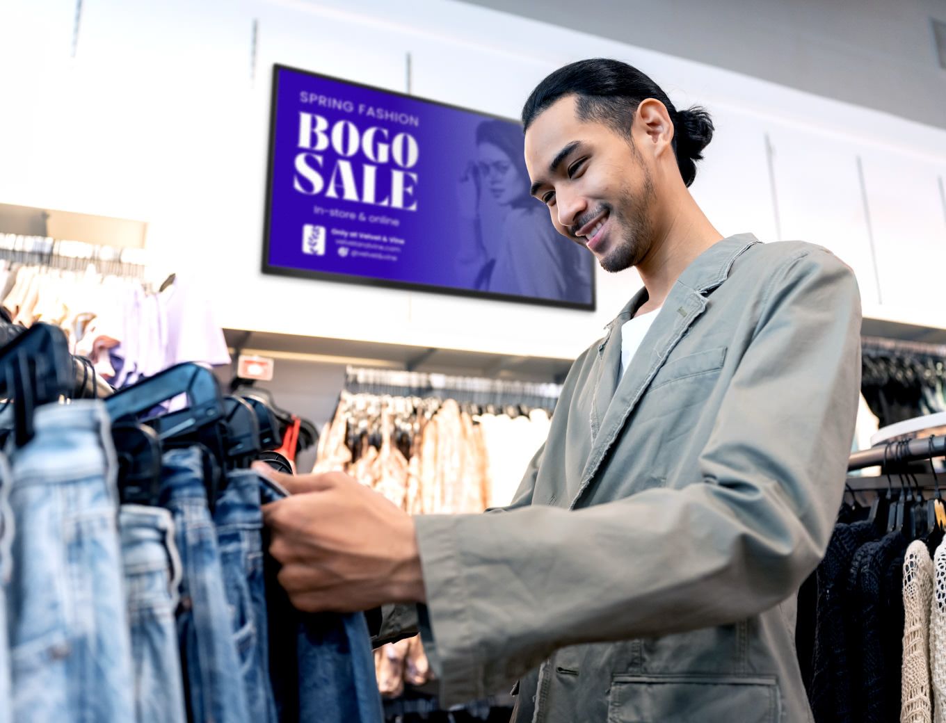Man browsing through a rack of pants in a clothing store with a slightly blurred tv behind him with a sales promotion ad displayed