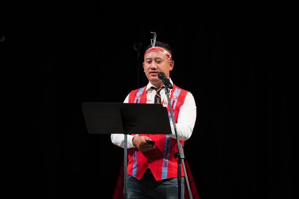 Photograph of a speaker in traditional attire addressing the audience at the First Naga Baptist Fundraising Cultural Event.