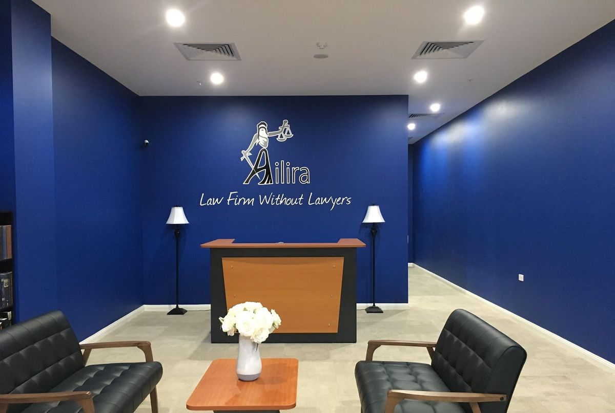 The reception of Ailira's law firm without lawyers: a deep blue room with the Ailira logo and the line "Law Firm Without Lawyers" on the wall above an empty reception desk, flanked by two floor lamps and two black leather armchairs