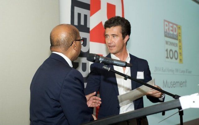 Musement's co-founder accepting a Red Herring Top 100 award on stage, the company name on the screen behind him