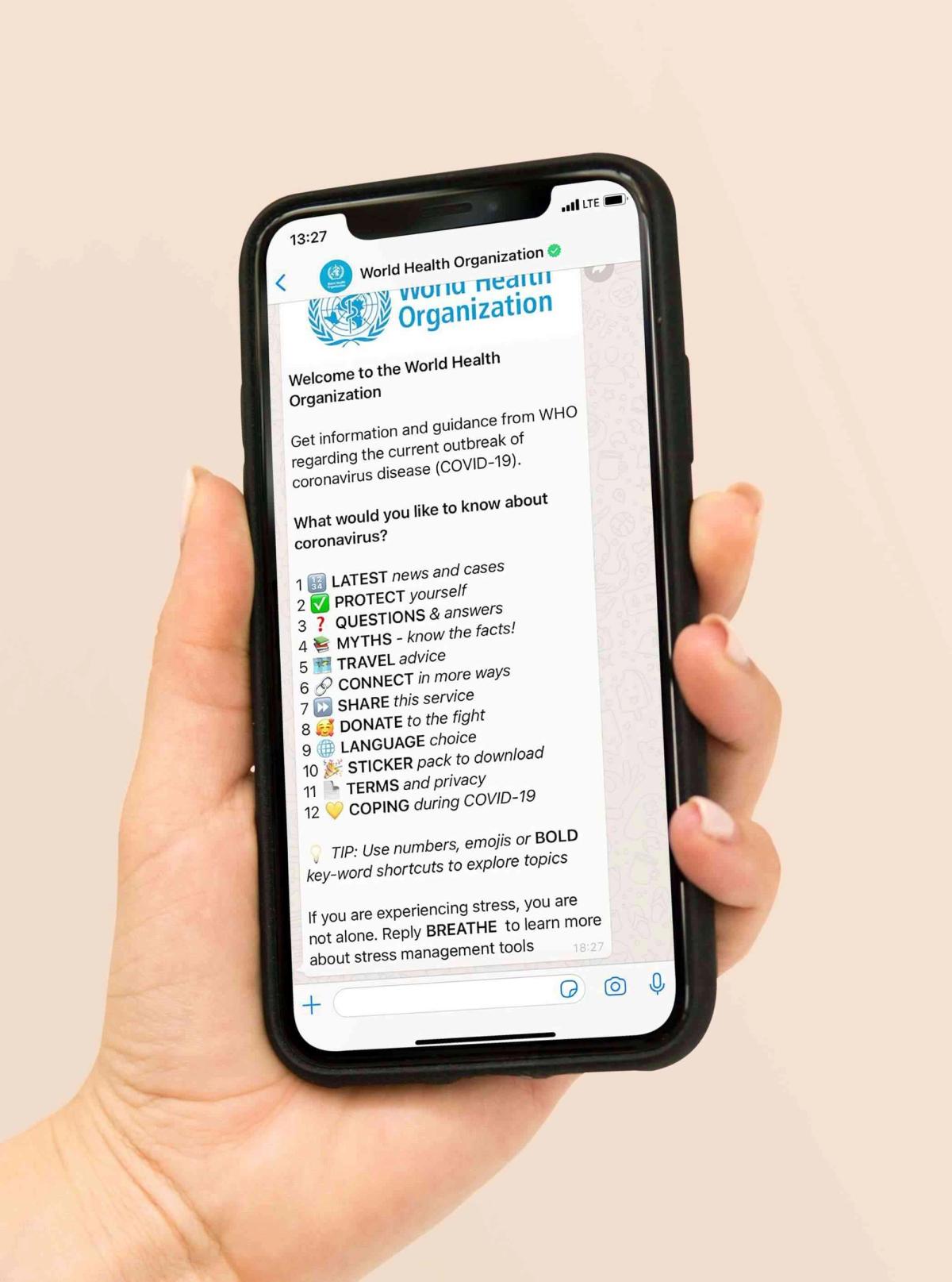 A hand holding a phone on the World Health Organization's verified WhatsApp chatbot: a welcome message offering a numbered menu of twelve coronavirus topics - latest news and cases, protect yourself, questions and answers, myths, travel advice, connect, share, donate, language choice, sticker pack, terms and privacy, coping during COVID-19