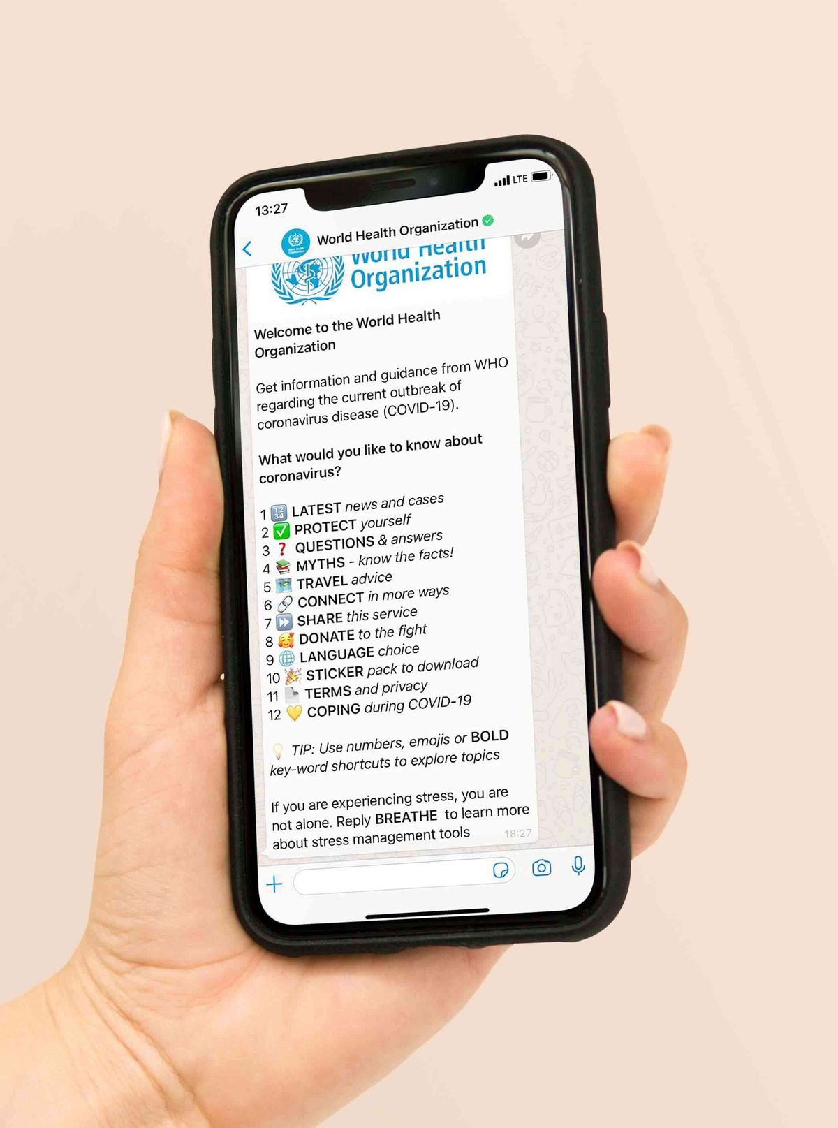 A hand holding a phone on the World Health Organization's verified WhatsApp chatbot: a welcome message offering a numbered menu of twelve coronavirus topics - latest news and cases, protect yourself, questions and answers, myths, travel advice, connect, share, donate, language choice, sticker pack, terms and privacy, coping during COVID-19
