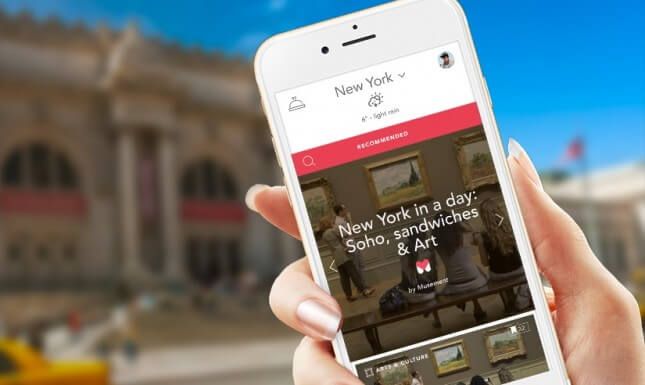 A hand holding an iPhone showing the Musement app's New York city guide, with a “New York in a day: Soho, sandwiches & Art” recommendation card, in front of a museum facade