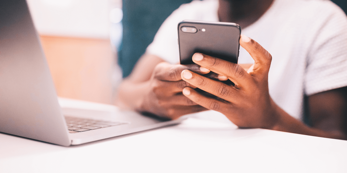 Close-up of a person in a white T-shirt holding a smartphone in both hands at a desk, beside an open laptop
