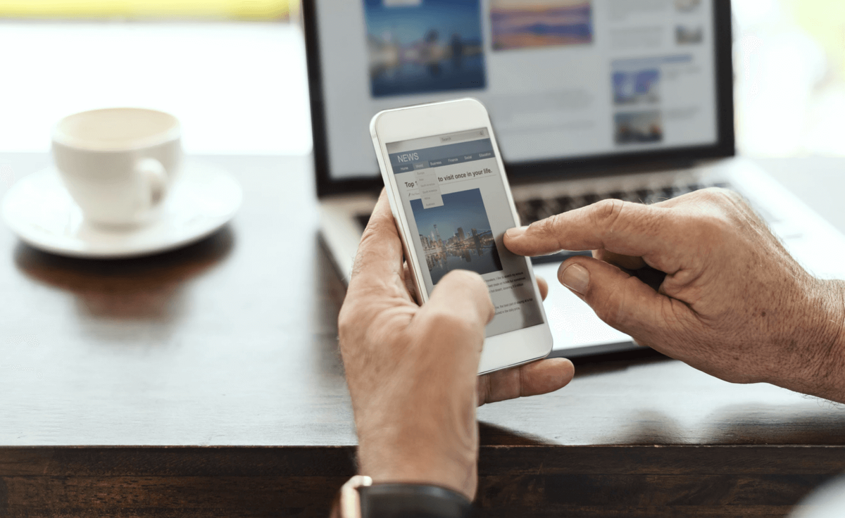 A reader browsing a news site on a phone with the same site open on a laptop behind