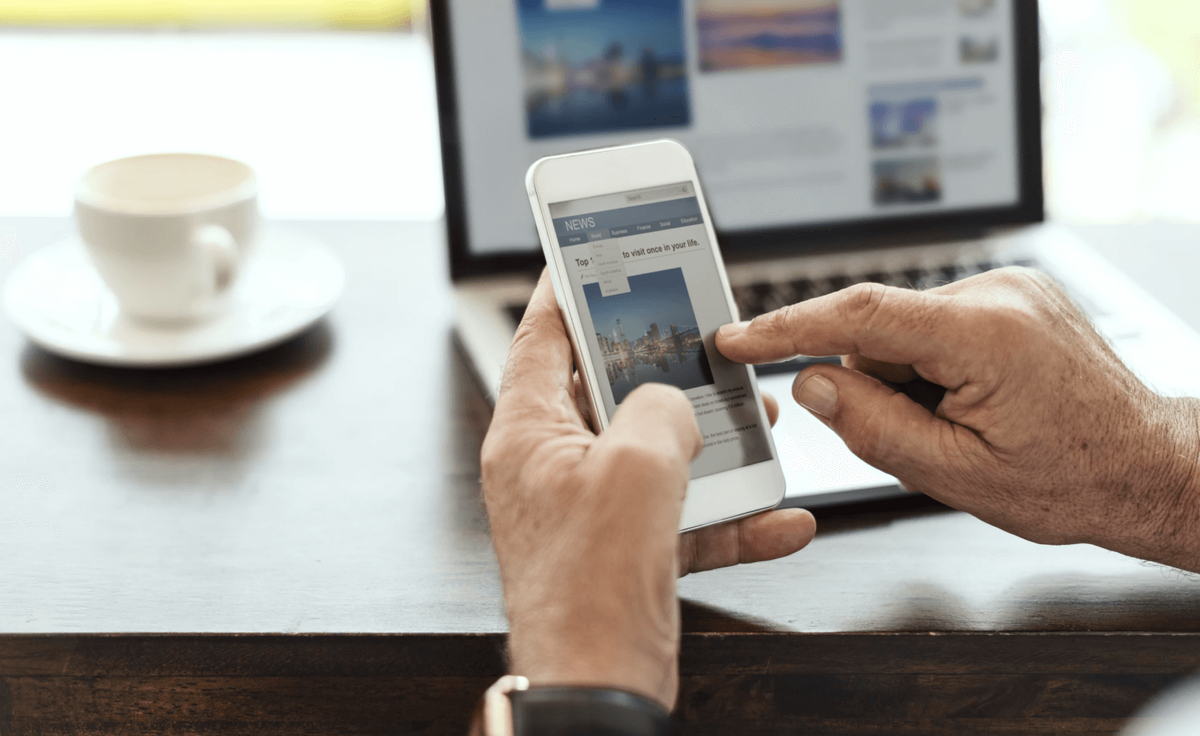 A reader browsing a news site on a phone with the same site open on a laptop behind