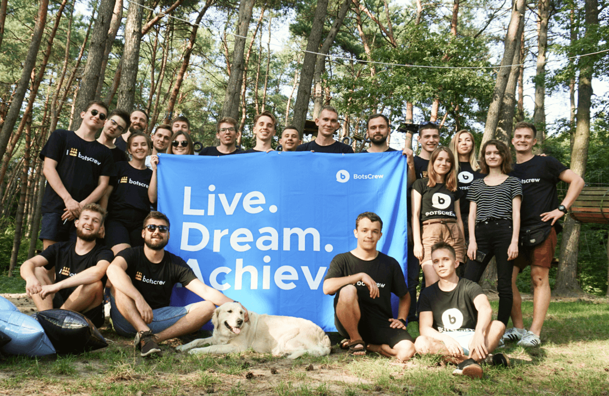The BotsCrew team photographed outdoors in a pine forest behind a blue banner reading 'Live. Dream. Achieve.'