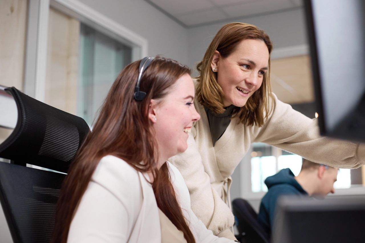 Twee collega's met headset kijken lachend samen naar een computerscherm.