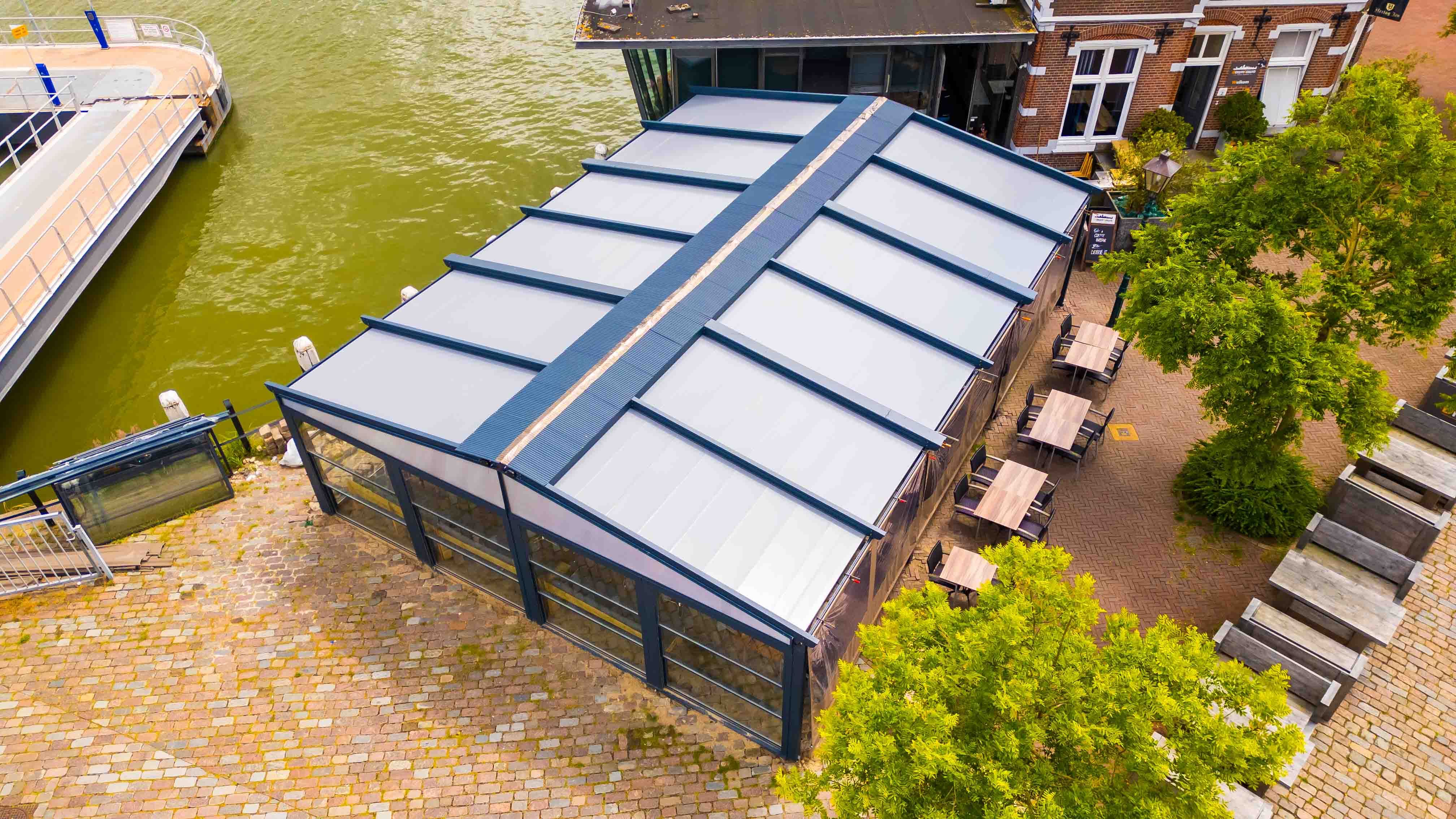 Schuine luchtfoto van een terrasoverkapping met gesloten dak aan het water, met een steiger links en tafels en een boom rechts, gemaakt door Bold Productions