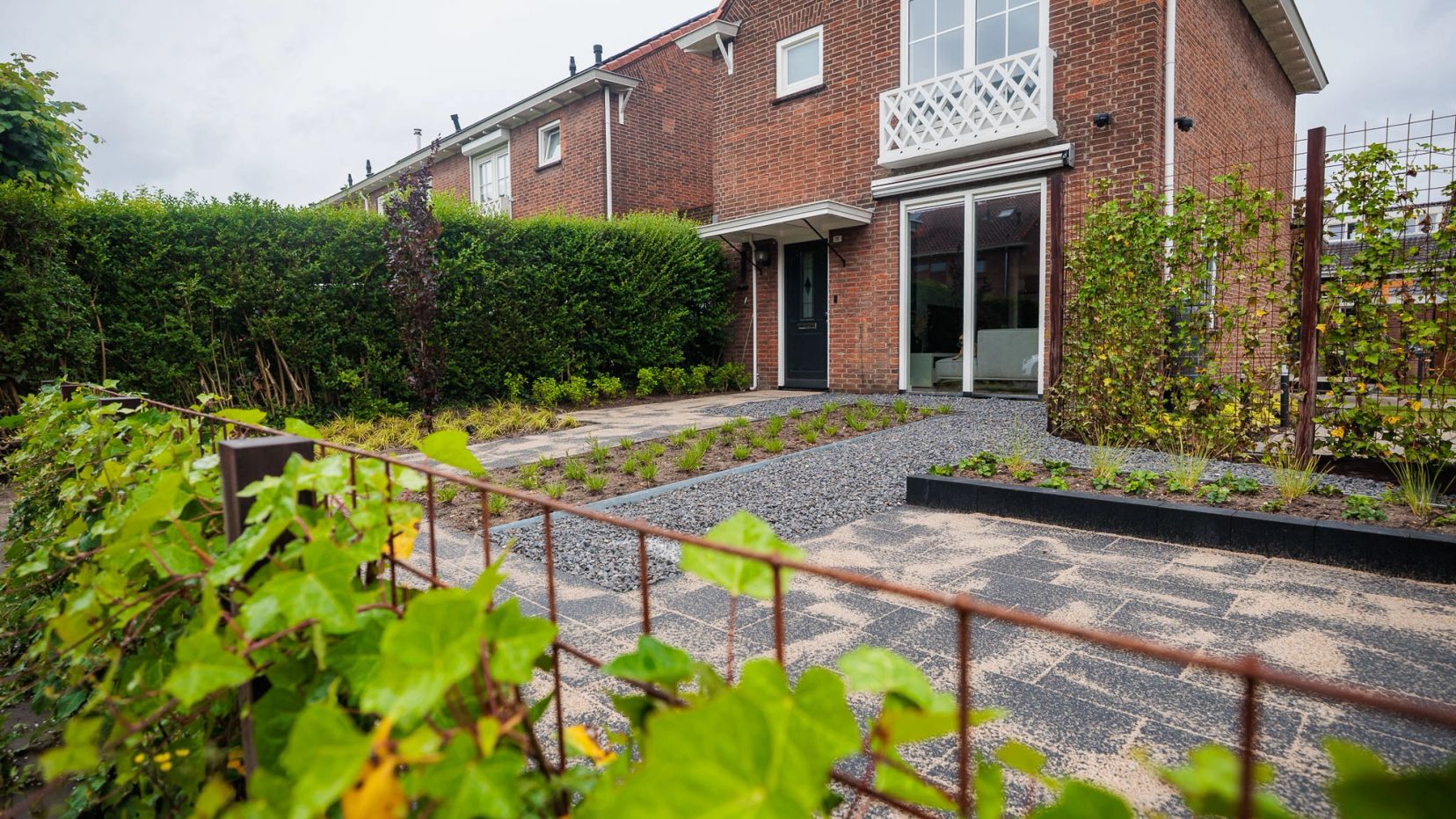 Voortuin met roestkleurige klimframes, haag en klinkers voor een rijwoning.