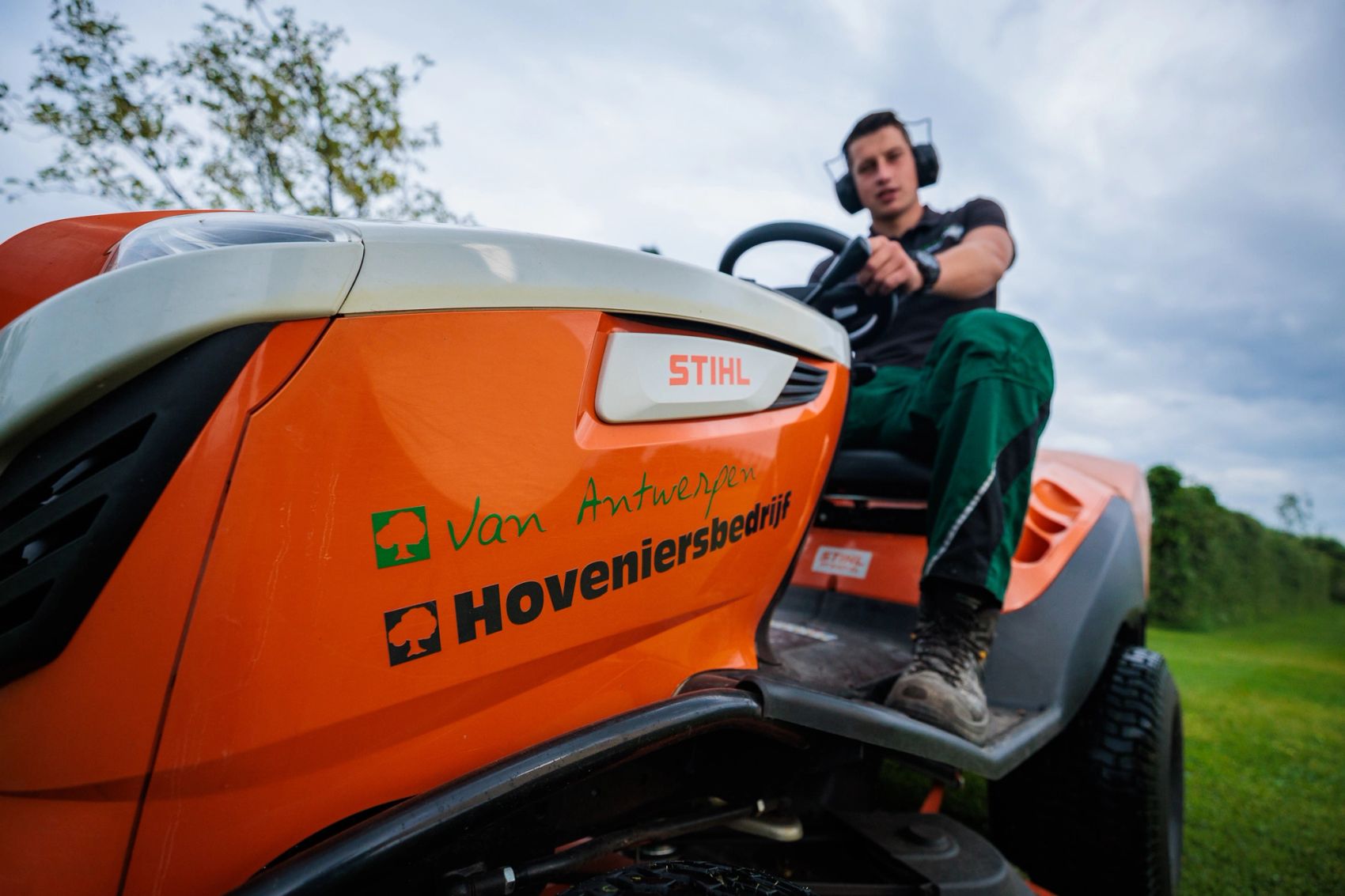 Close-up van een oranje zitmaaier met het logo van Hoveniersbedrijf Van Antwerpen en hovenier achter het stuur, gemaakt door Bold Productions