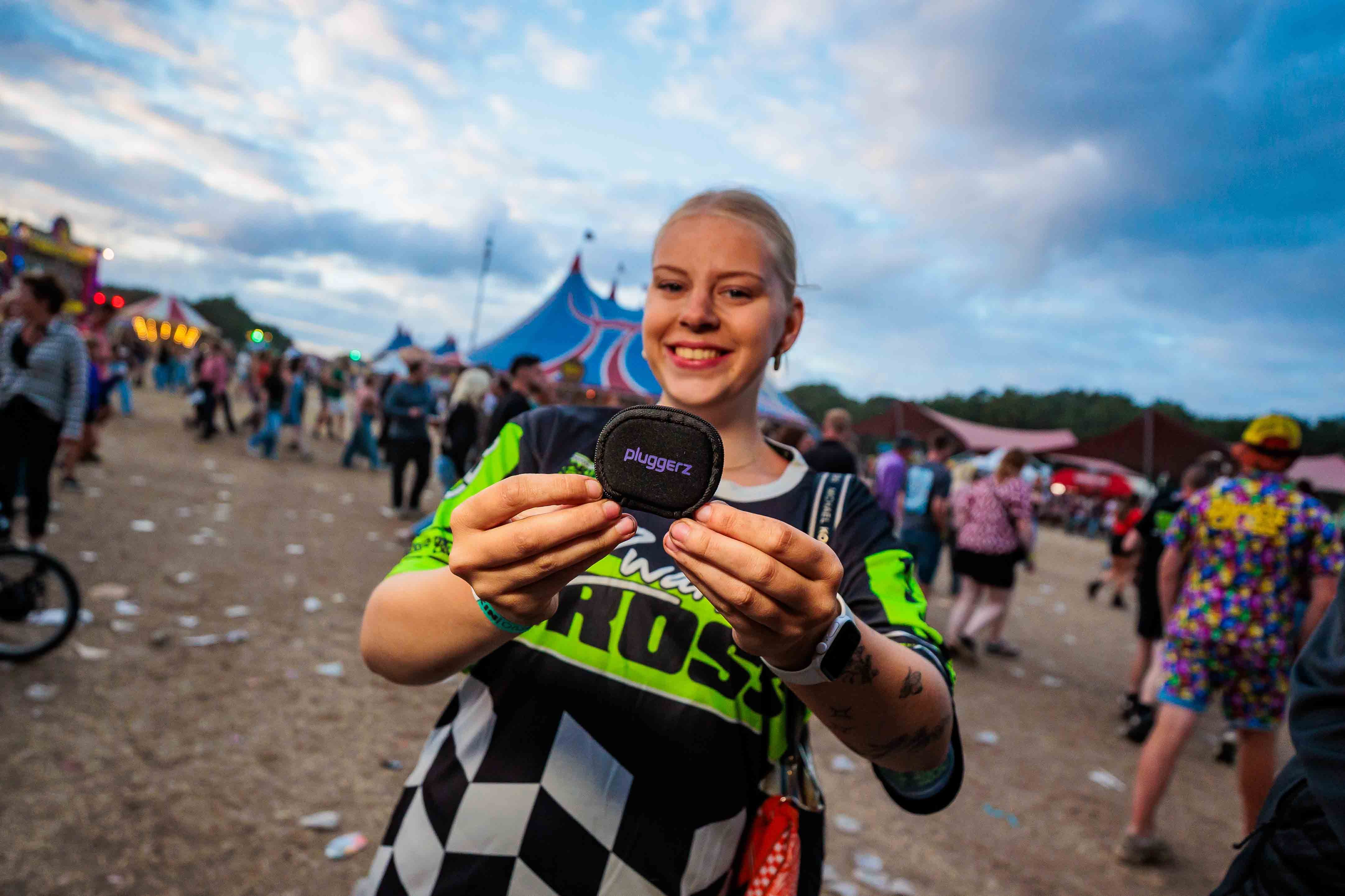 Jonge vrouw in een zwart-groen festivalshirt met een geblokte rok die een Pluggerz-etui vasthoudt op het festivalterrein bij avondlicht, gemaakt door Bold Productions
