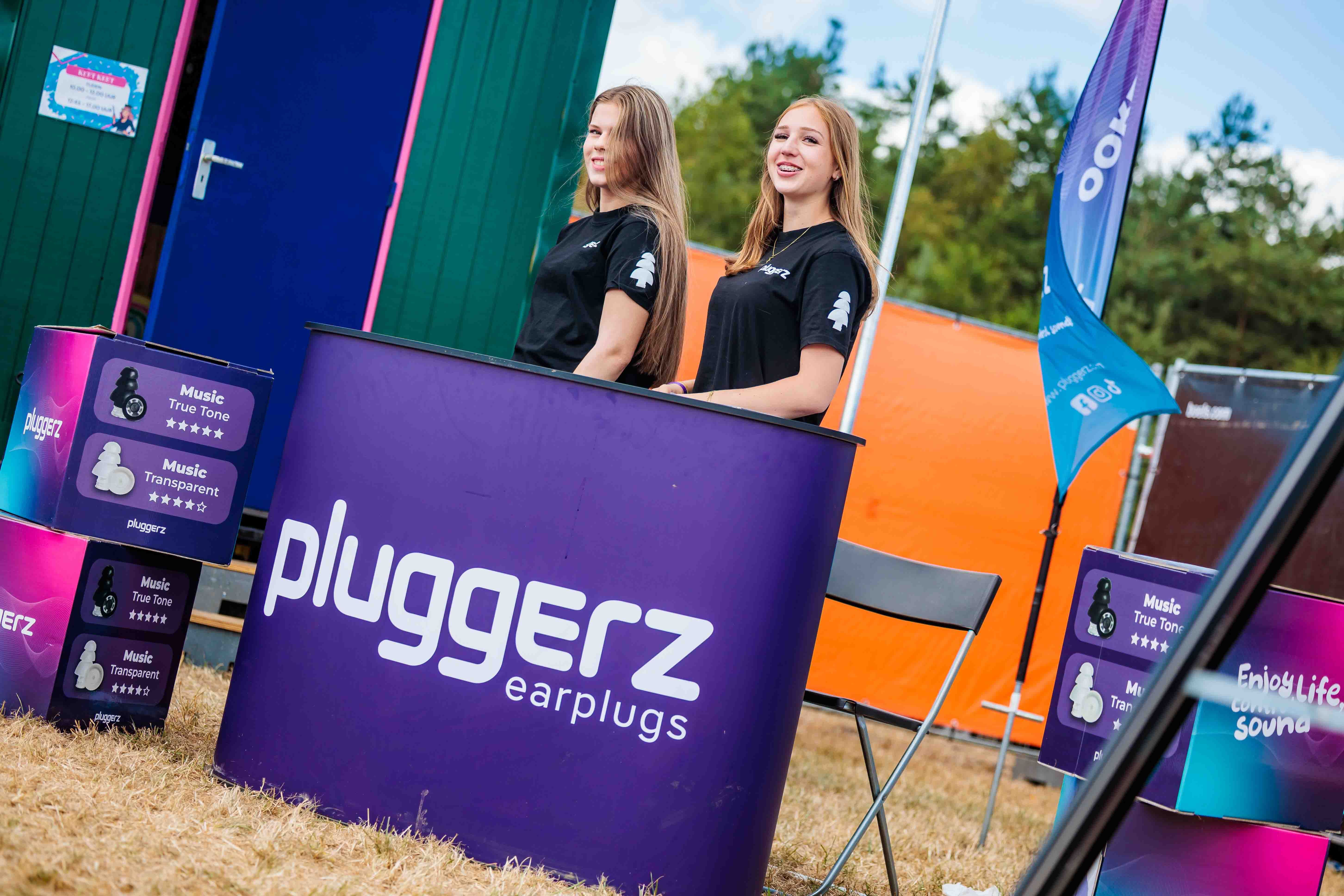 Twee medewerkers in zwarte shirts achter een paarse Pluggerz earplugs-balie naast een groene container, gemaakt door Bold Productions