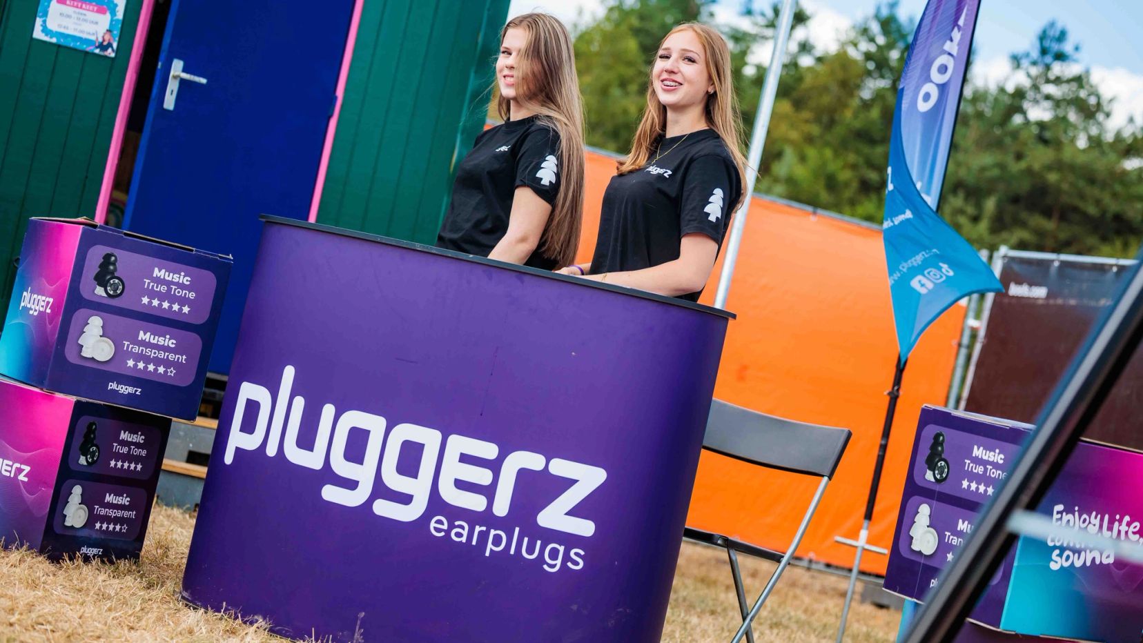 Twee medewerkers in zwarte shirts achter een paarse Pluggerz earplugs-balie naast een groene container, gemaakt door Bold Productions