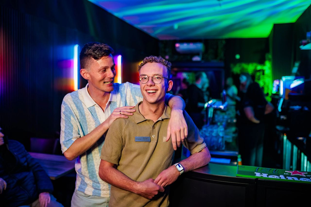 Twee gastheren lachen samen onder kleurrijke plafondverlichting bij Timestamp Arnhem.