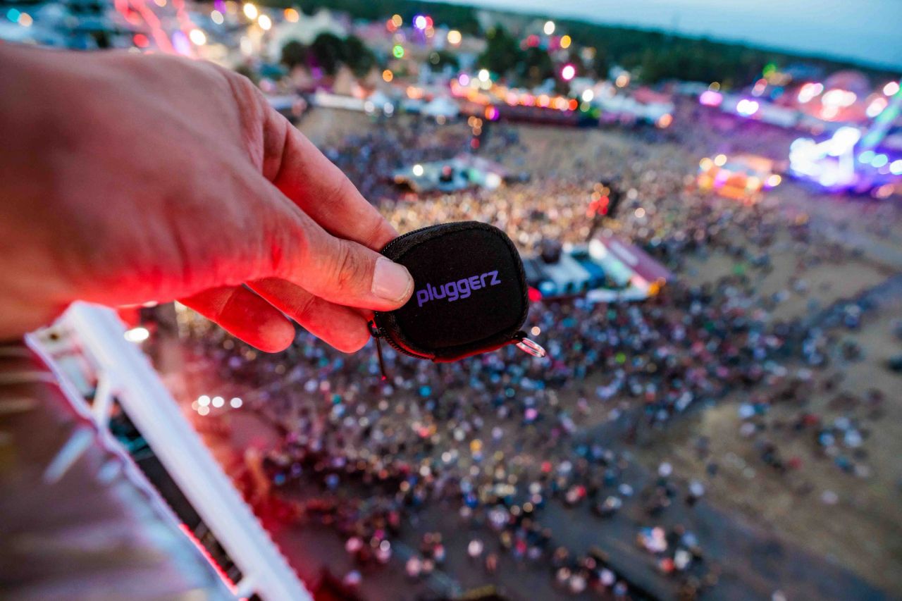 Hand met een zwart Pluggerz-etui op grote hoogte, met het drukke festivalterrein diep eronder, gemaakt door Bold Productions