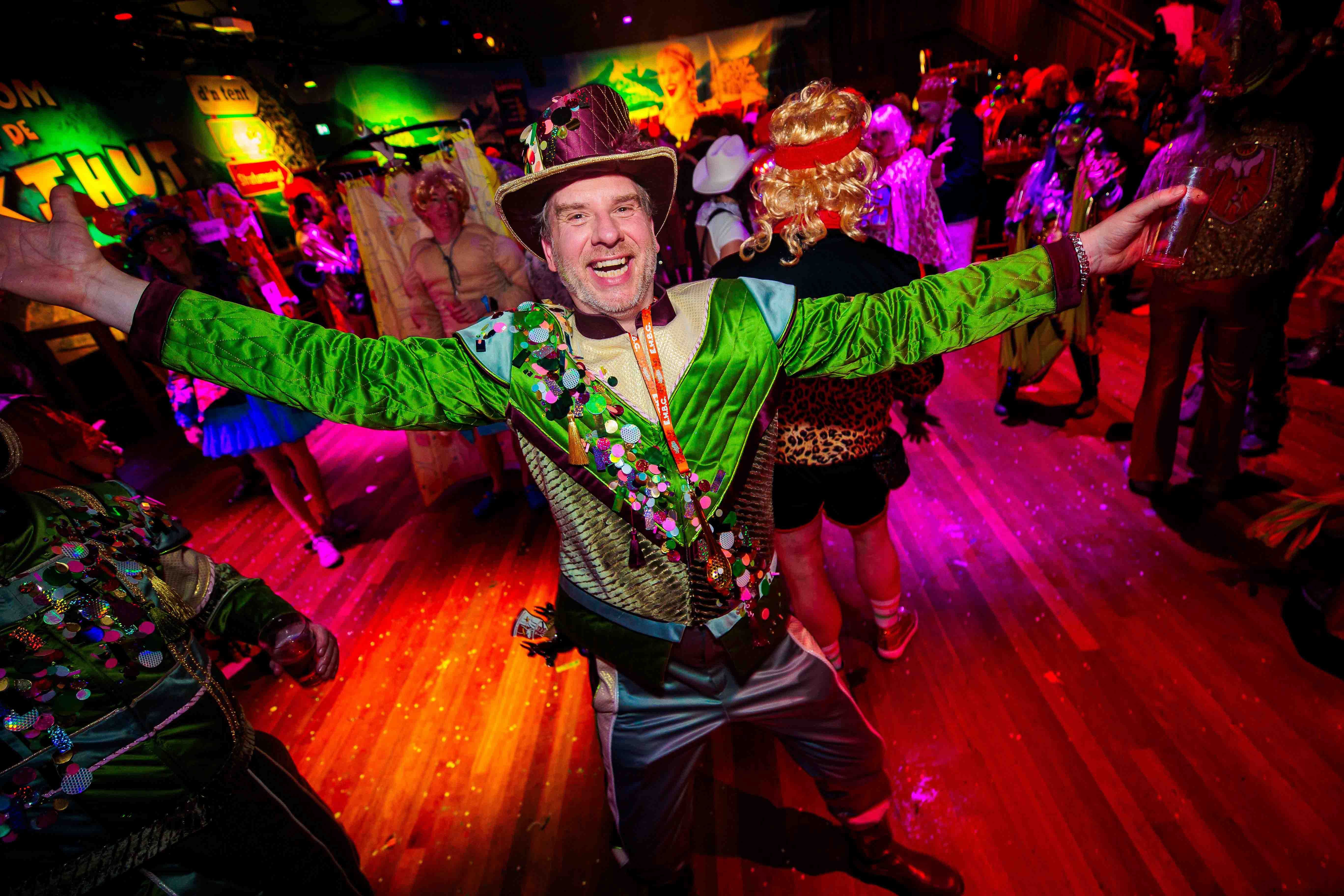 Ralph Buisman in een kleurrijk feestoutfit met uitgestrekte armen in een rood verlichte feestzaal