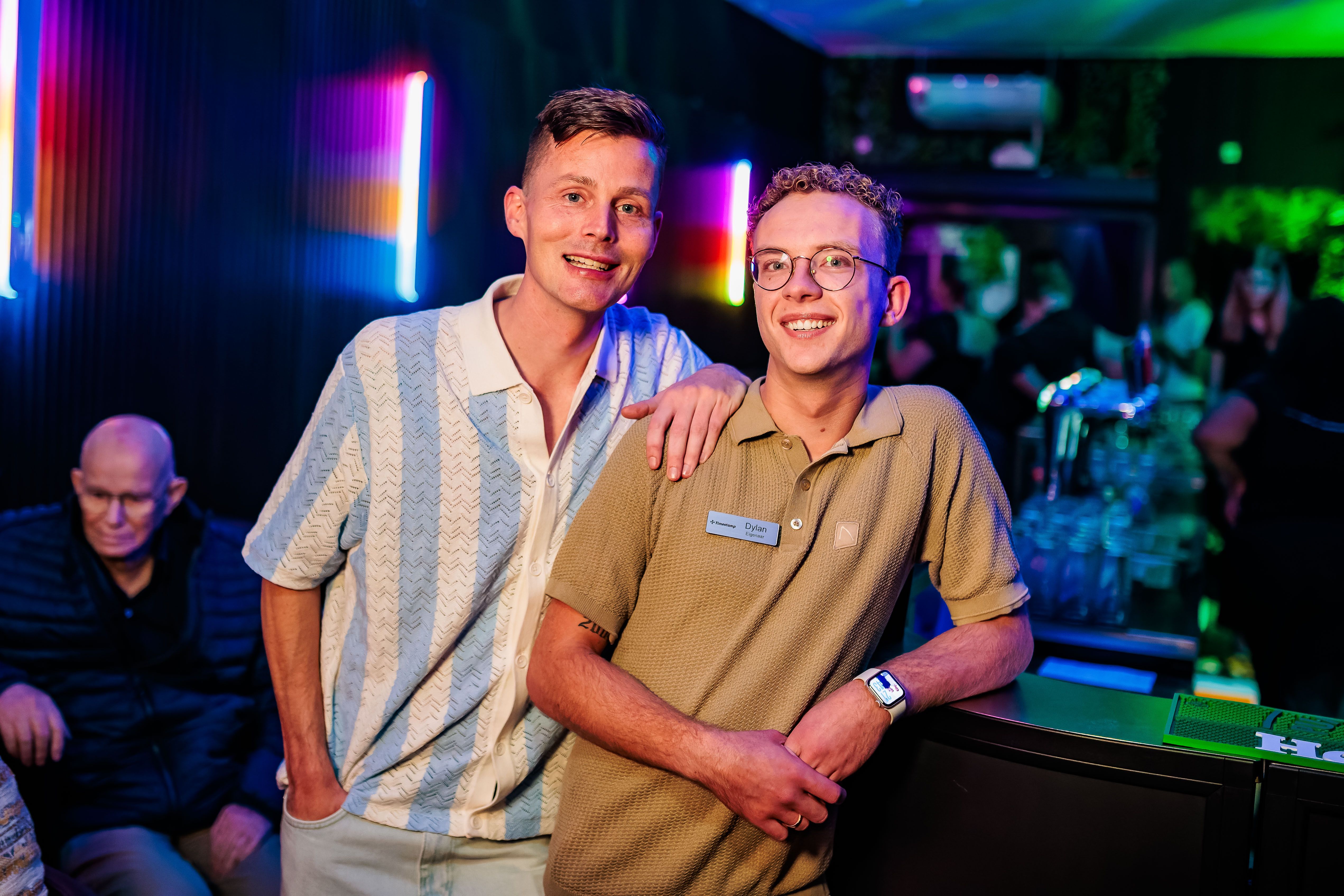 Twee gastheren in gestreept en beige overhemd poseren in de neonverlichte zaak, gemaakt door Bold Productions