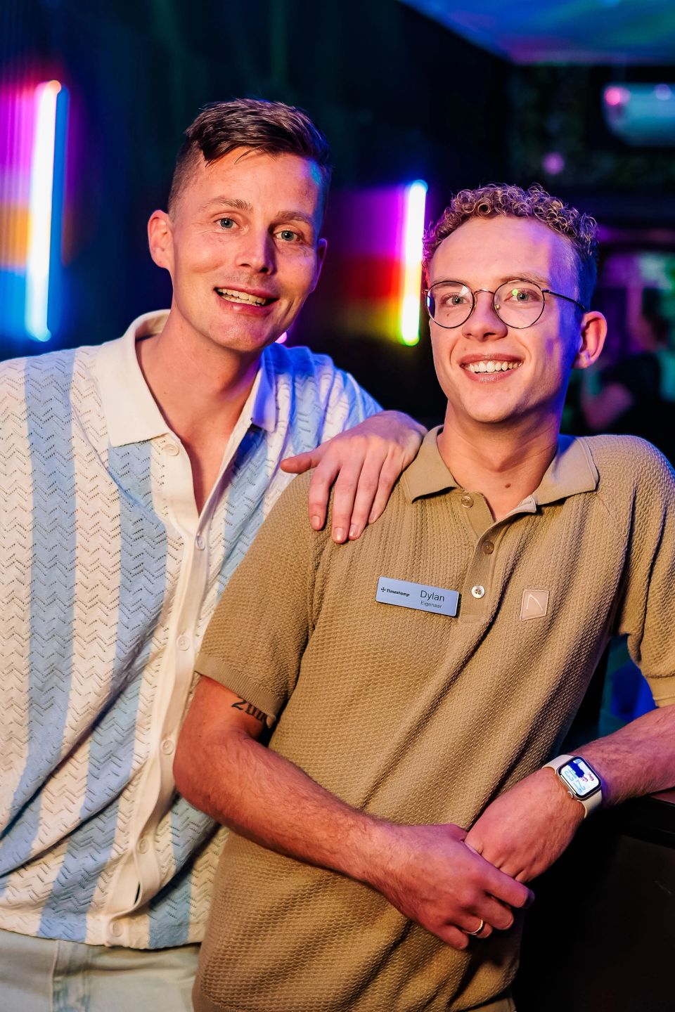 Twee gastheren in gestreept en beige overhemd poseren in de neonverlichte zaak, gemaakt door Bold Productions