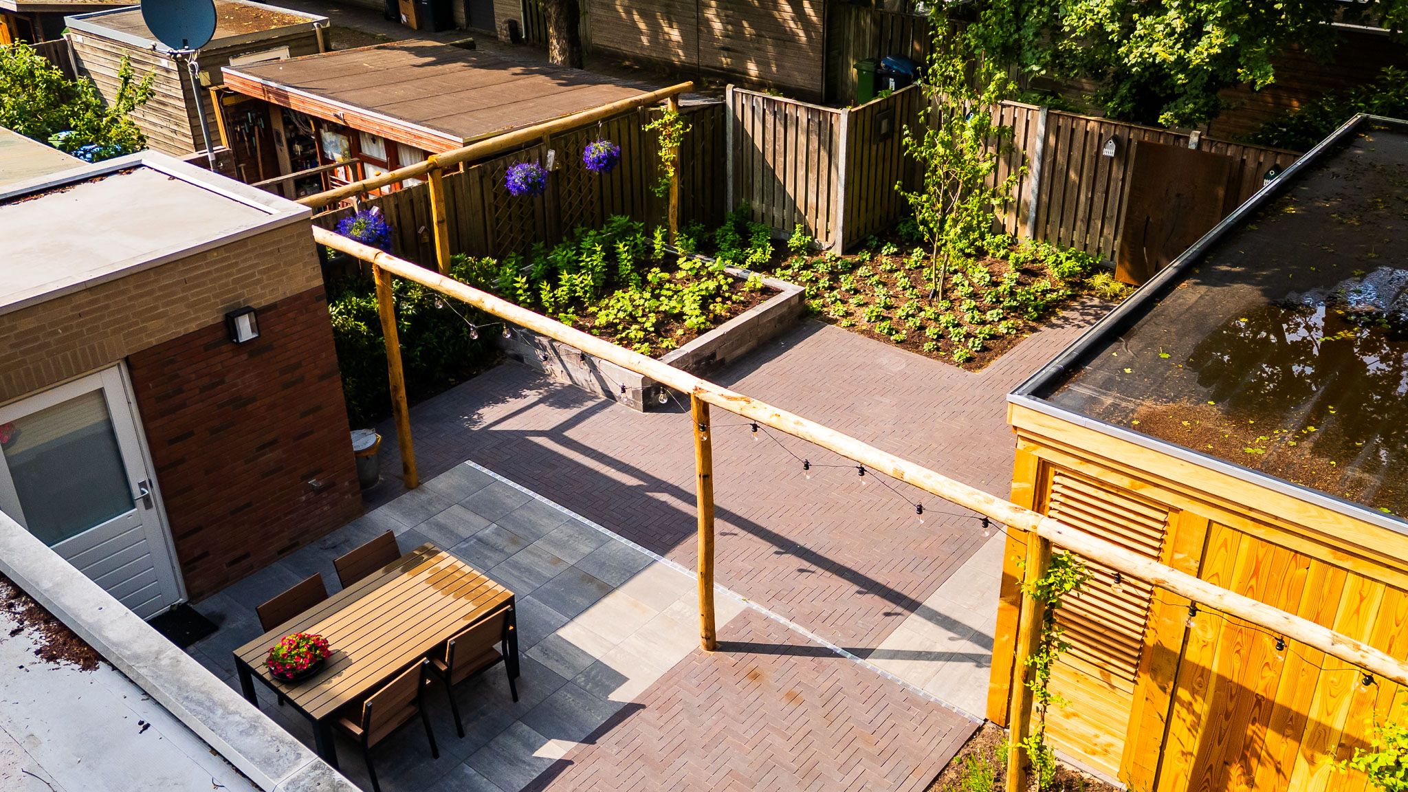Luchtfoto van een tuin met houten pergola, eettafel en groene borders.