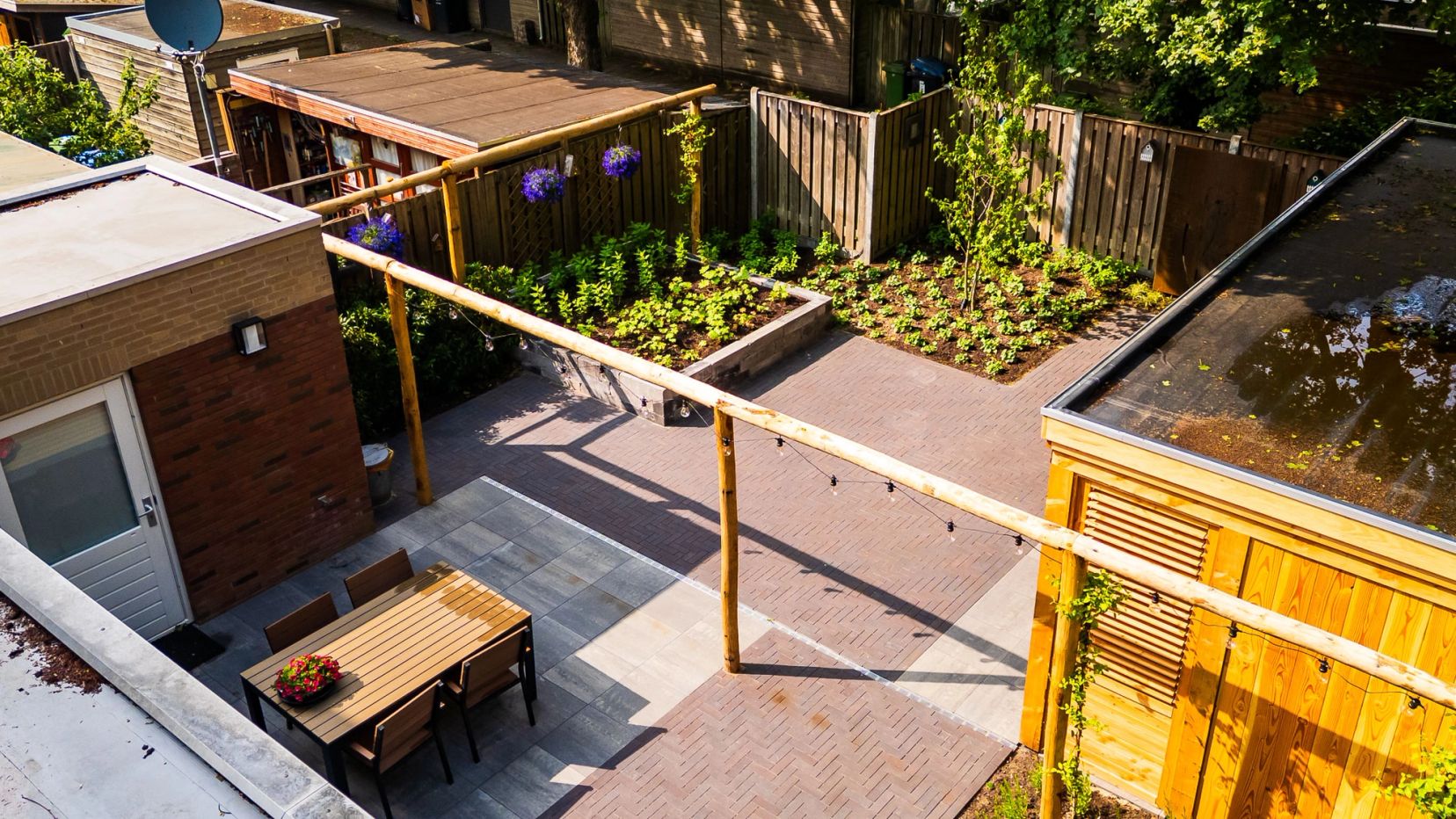 Luchtfoto van een tuin met houten pergola, eettafel en groene borders.