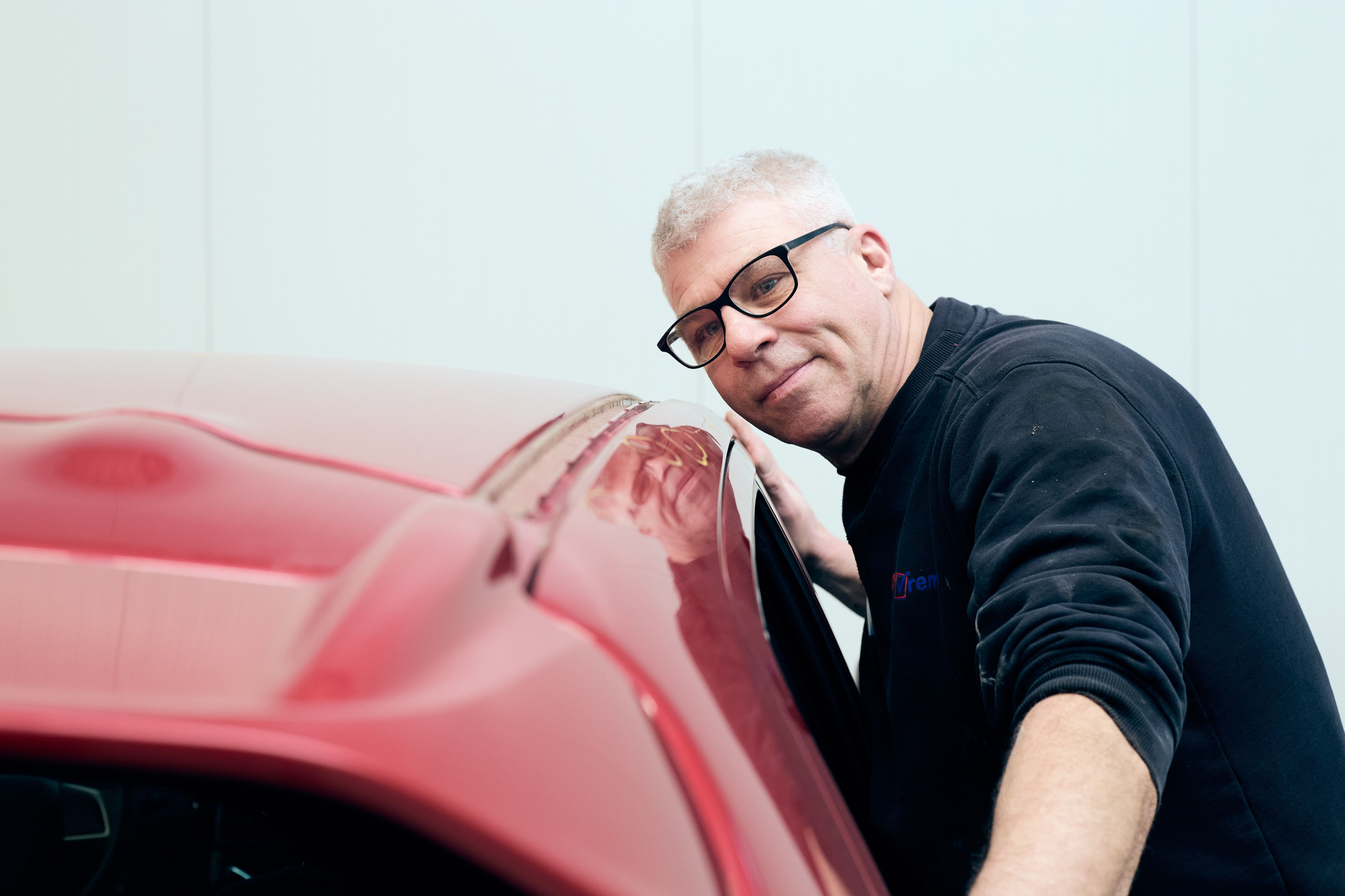 Lachende monteur met bril leunt tegen een rode auto bij Auto Vreman.