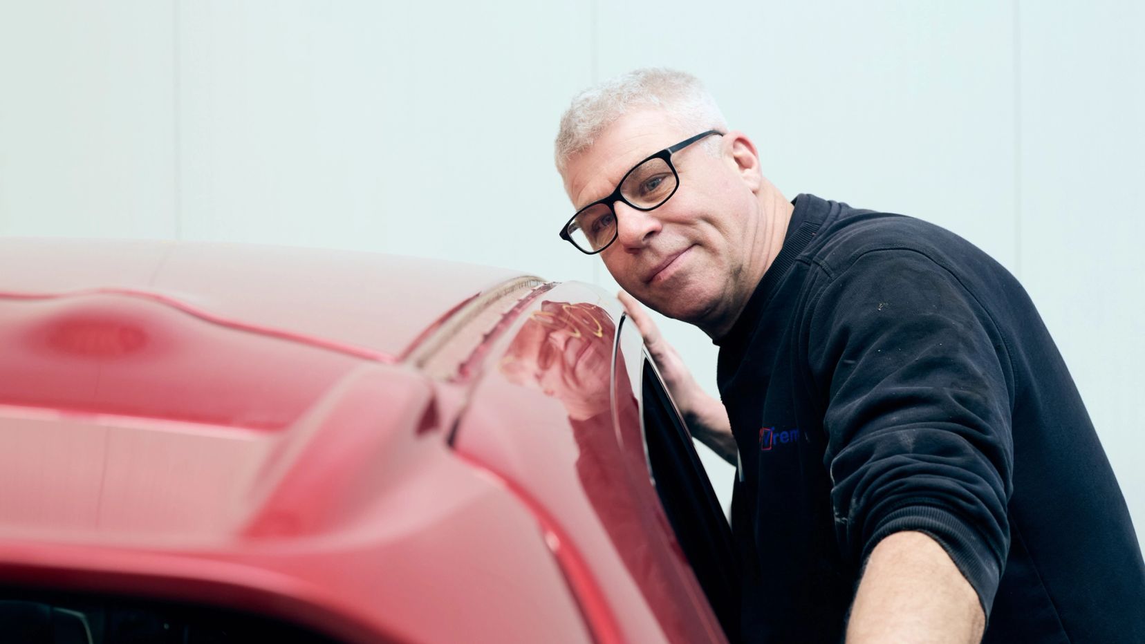 Lachende monteur met bril leunt tegen een rode auto bij Auto Vreman.