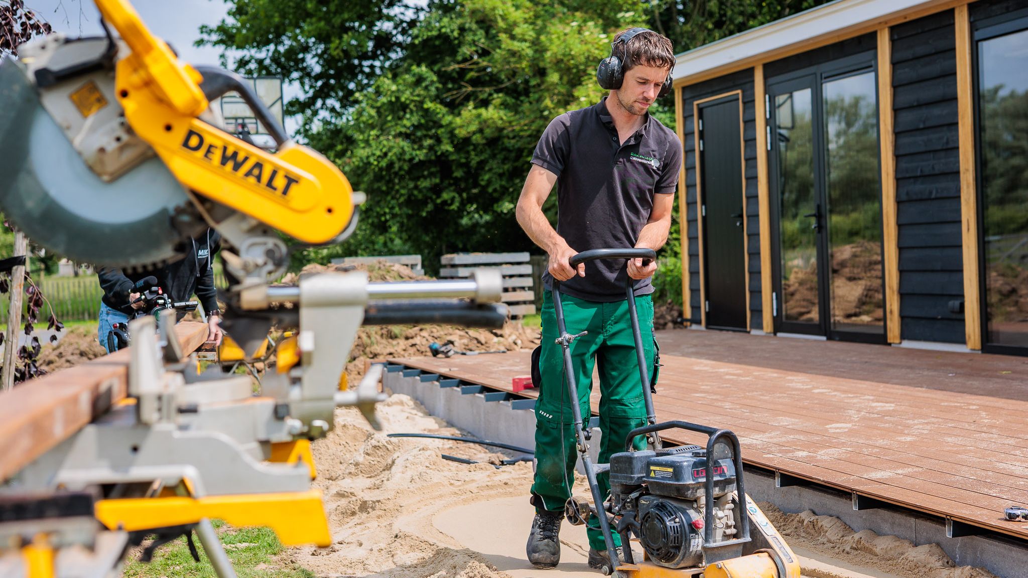 Hovenier werkt met een trilplaat naast een afkortzaag bij een houten tuinkamer, gemaakt door Bold Productions
