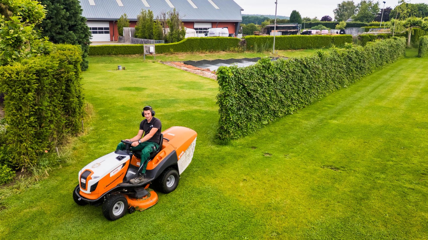 Hovenier maait een groot grasveld met een oranje zitmaaier langs een lange haag, gemaakt door Bold Productions