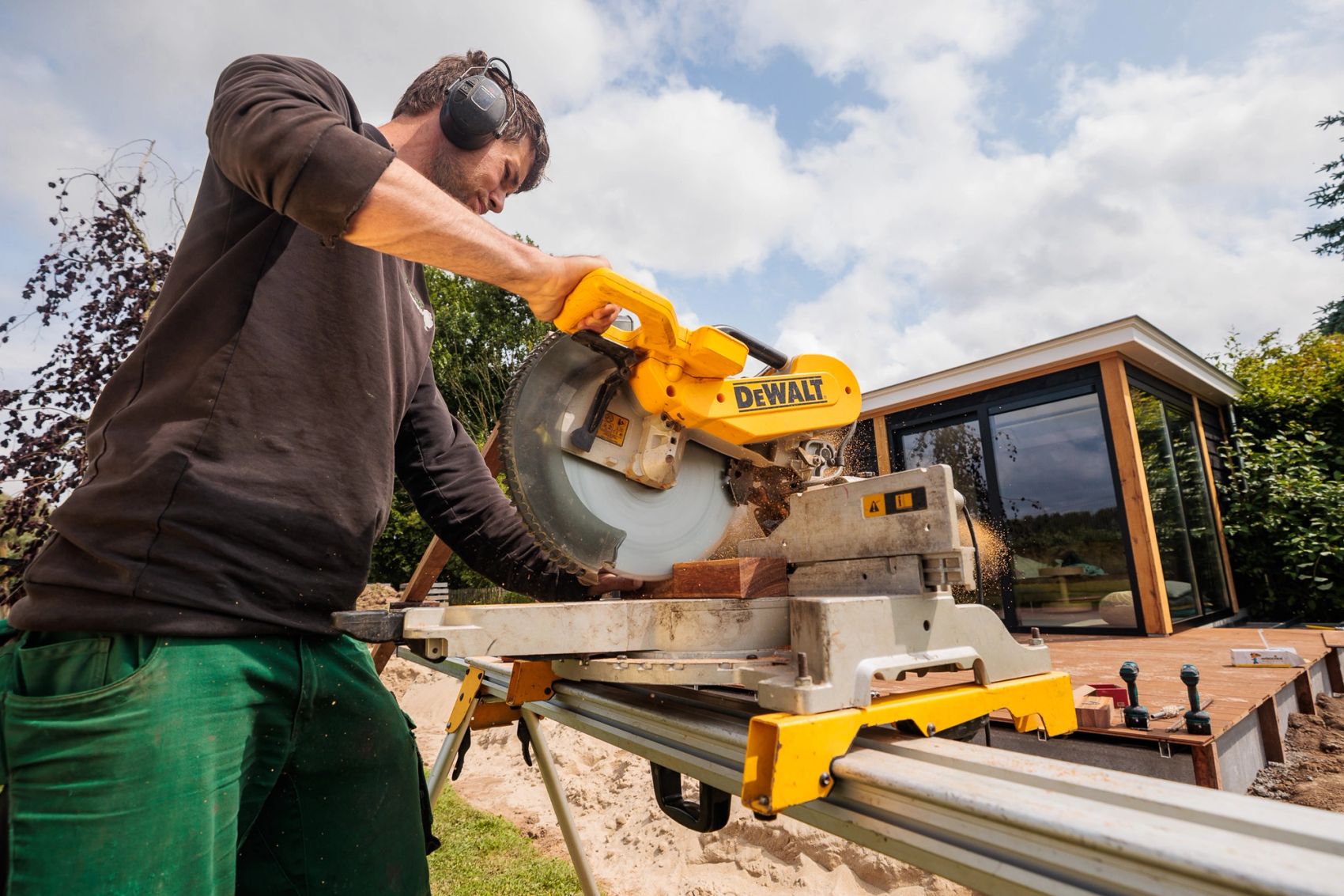 Hovenier met gehoorbescherming zaagt met een afkortzaag voor een houten tuinkamer, gemaakt door Bold Productions
