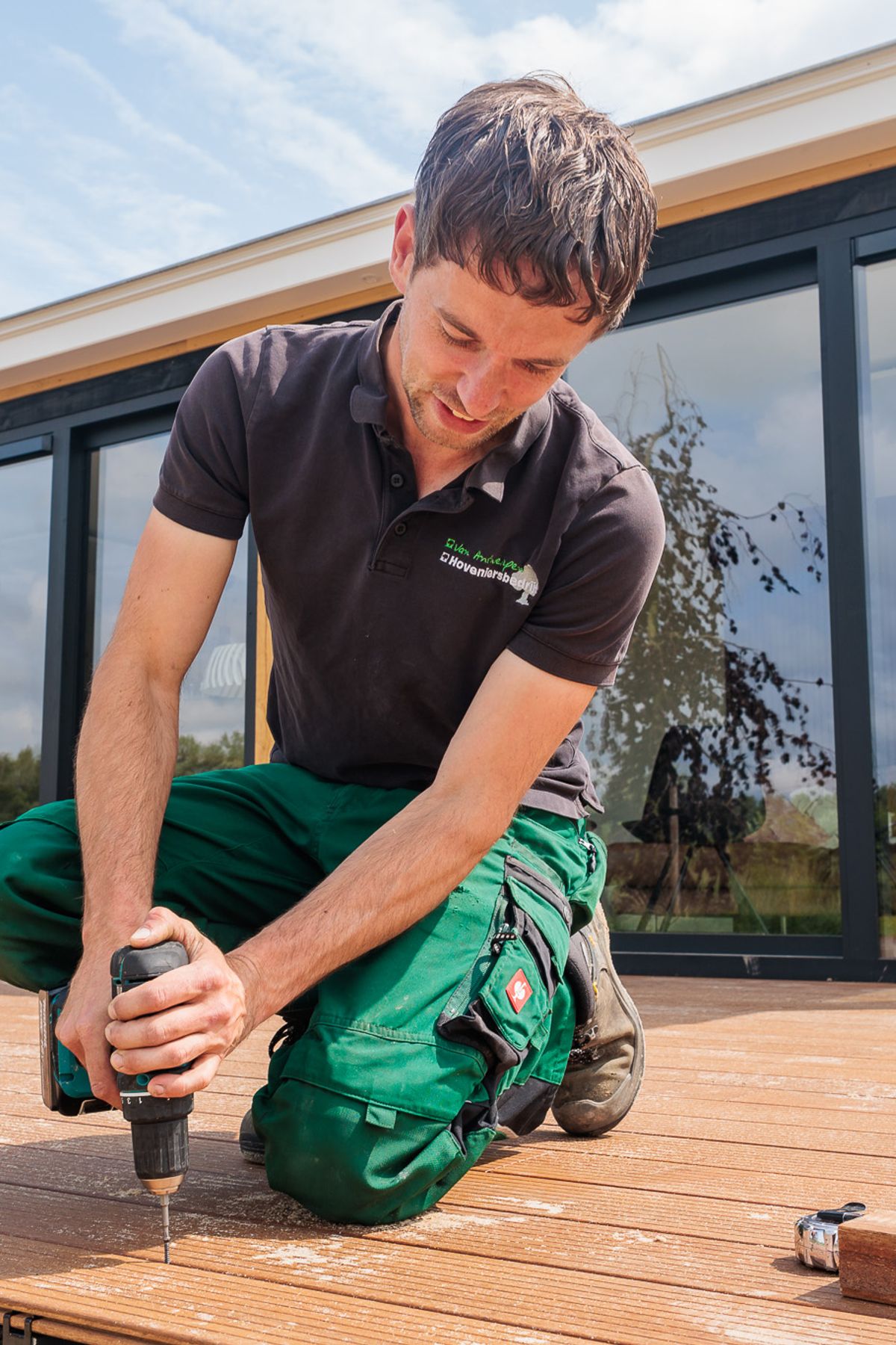 Hovenier knielt en schroeft een vlonderplank vast bij een houten tuinkamer, gemaakt door Bold Productions
