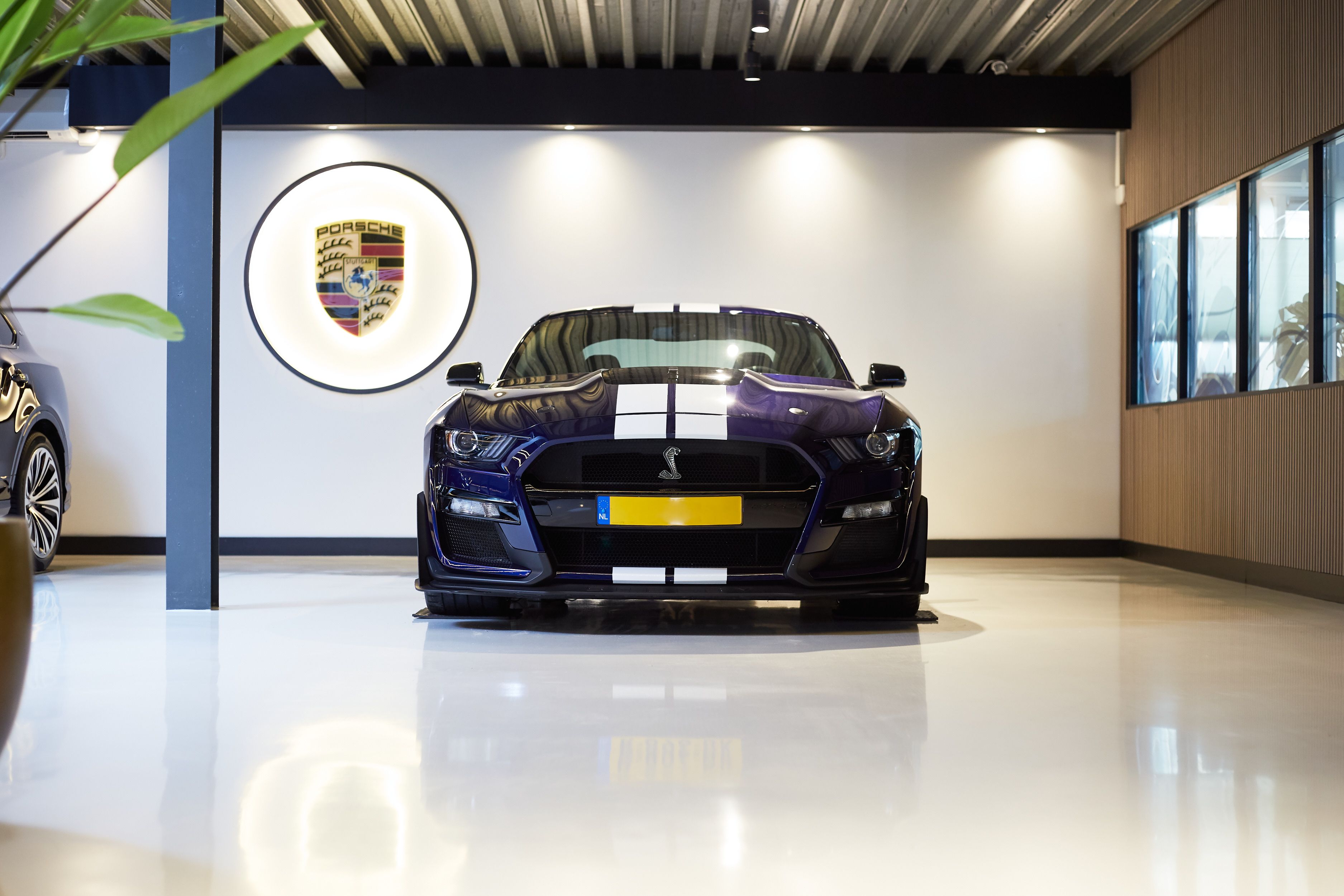 Blauwe sportauto met witte strepen onder een Porsche-logo in de showroom.