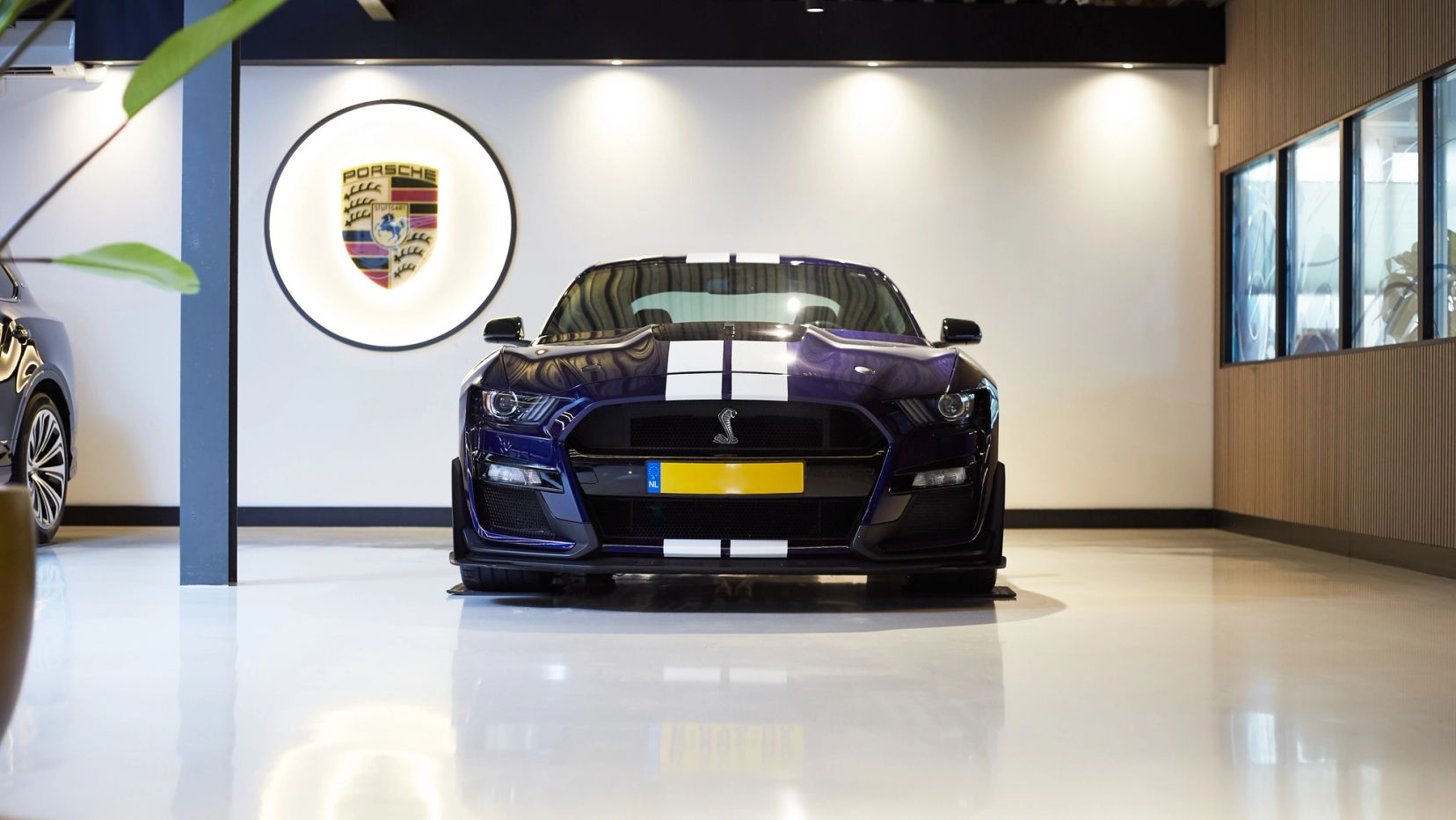 Blauwe sportauto met witte strepen onder een Porsche-logo in de showroom.