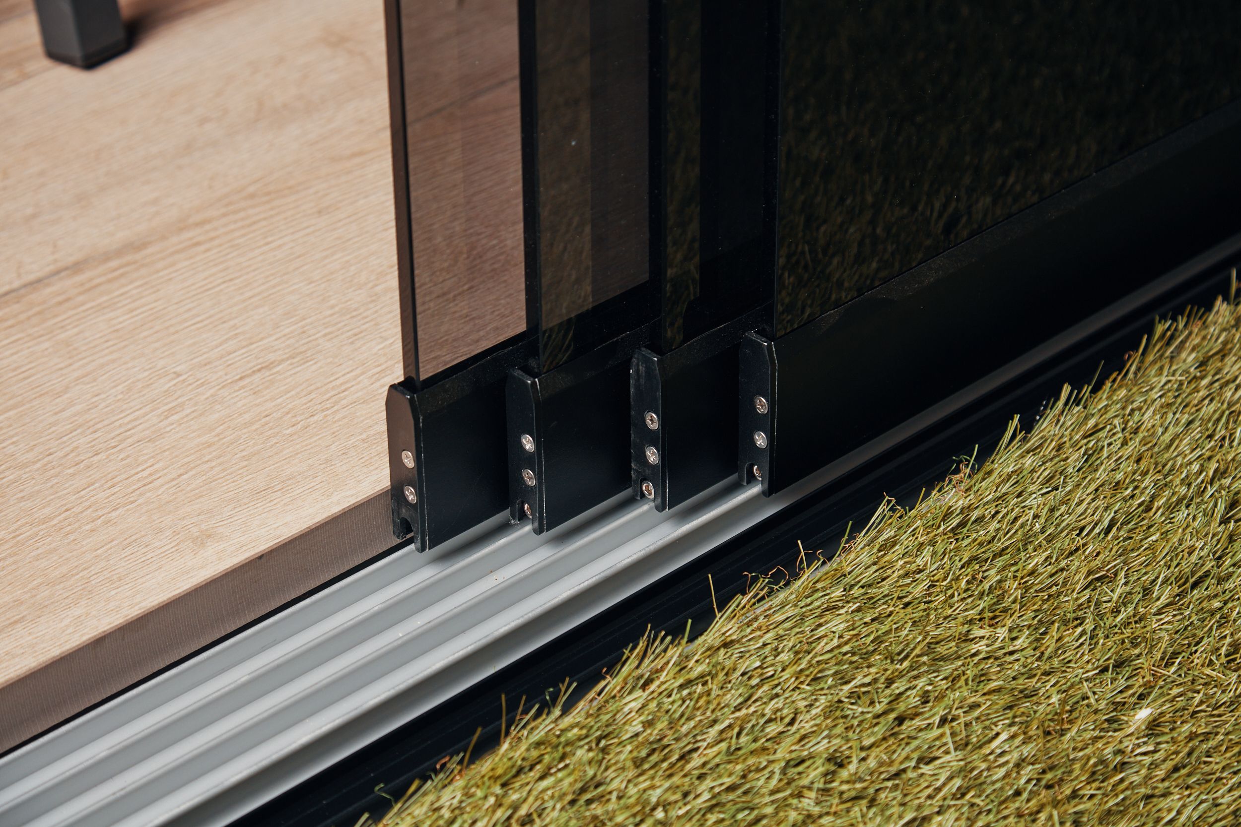 Detail van een glazen schuifwand op rails bij kunstgras.
