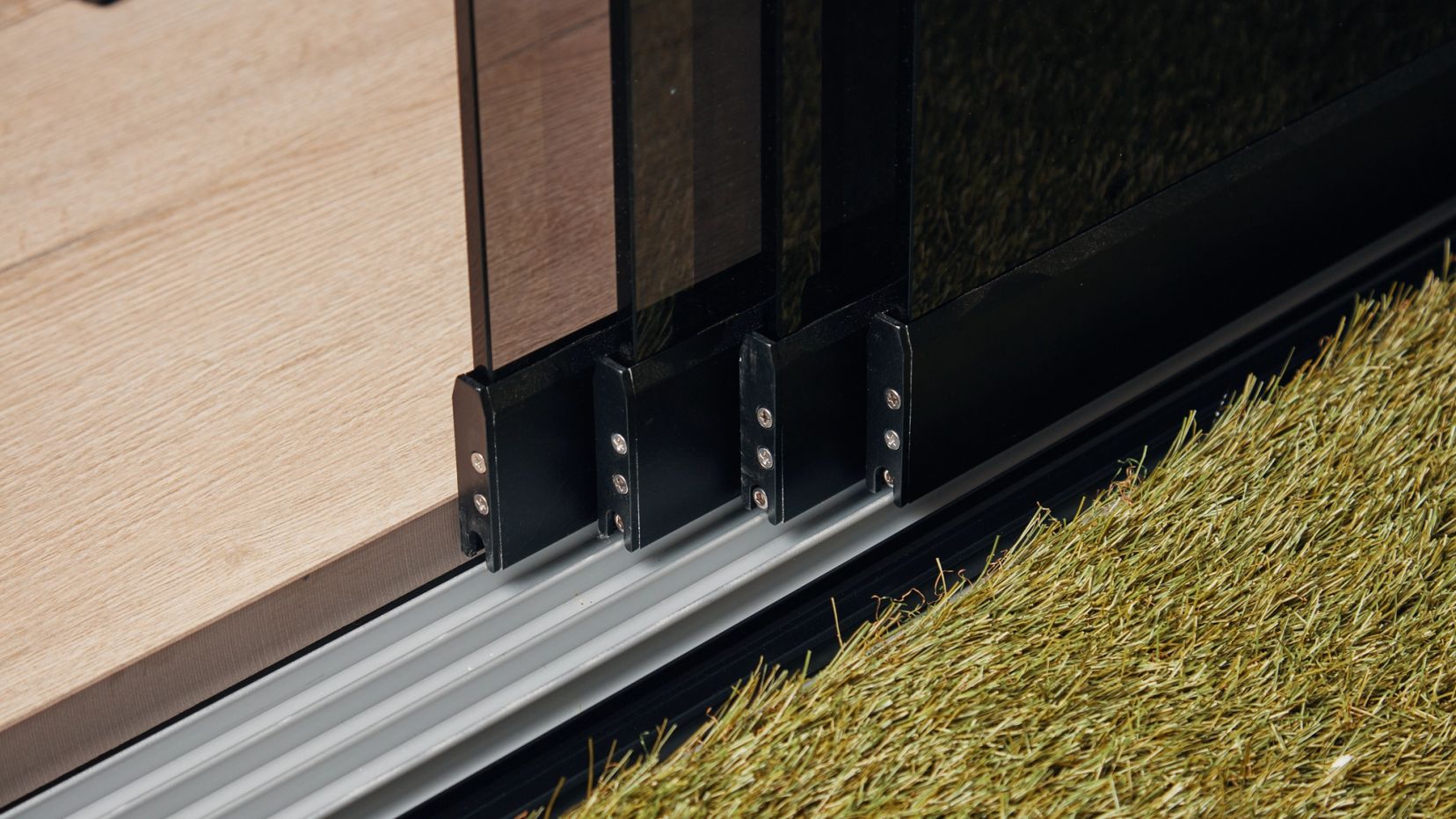 Detail van een glazen schuifwand op rails bij kunstgras.