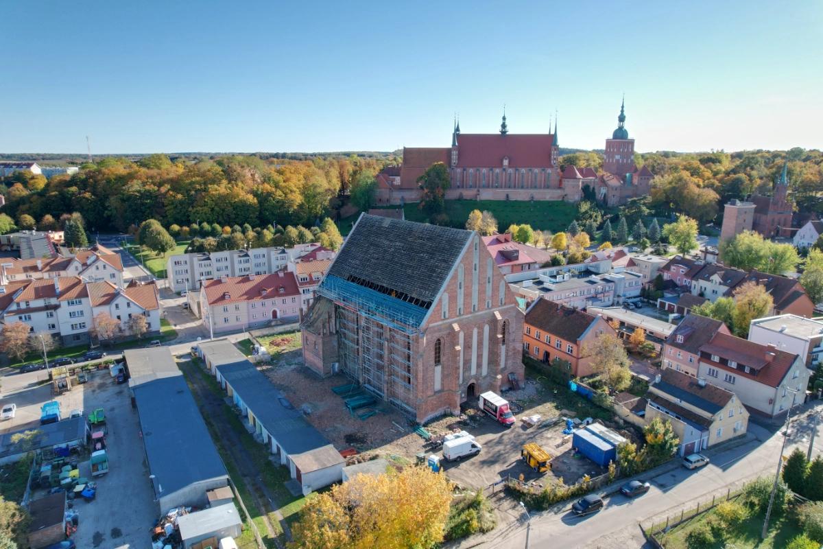 Widok z lotu ptaka na prace konserwatorsko-restauratorskie przy kościele św. Mikołaja we Fromborku, otoczonym zabudową miejską i katedrą na wzgórzu w tle.