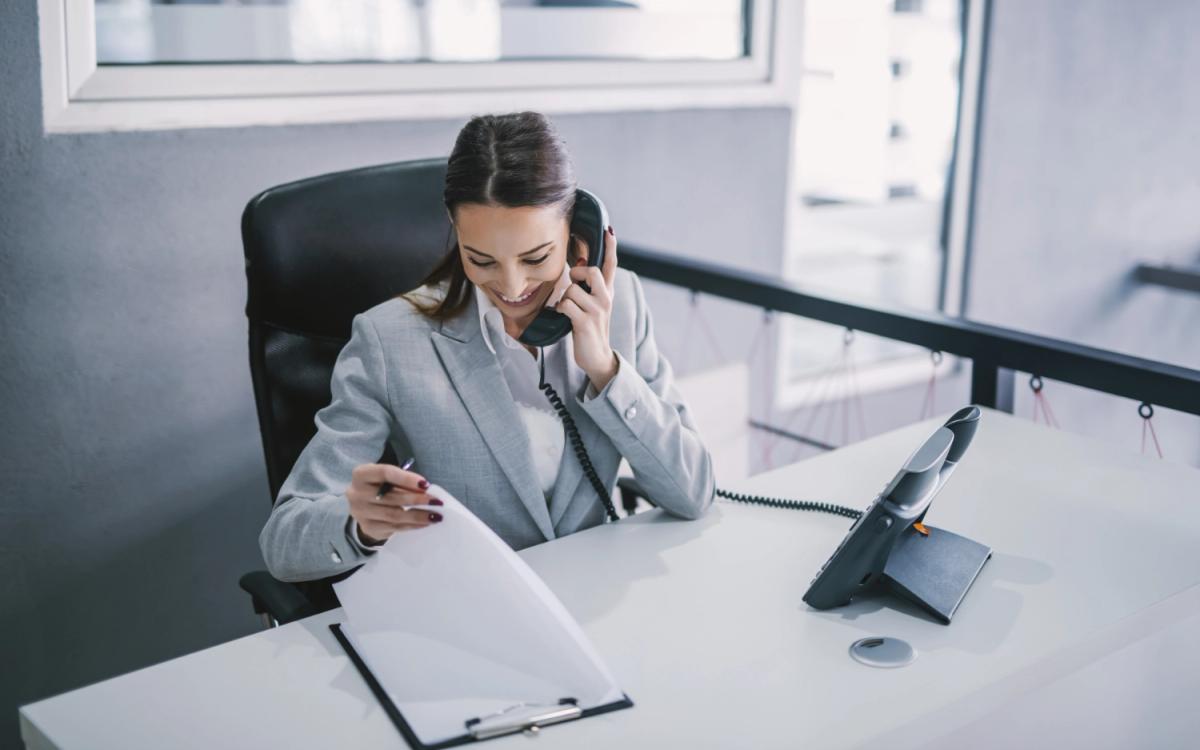 Pracowniczka biura w eleganckim garniturze rozmawia przez telefon i przegląda dokumenty, ilustrując wsparcie administracyjne i doradztwo w zakresie pozyskiwania dotacji.