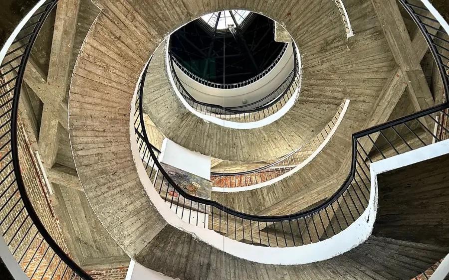 Widok w górę monumentalnej spiralnej klatki schodowej z betonu i cegły, ukazujący geometryczne formy i kunszt architektoniczny we wnętrzu zabytkowego budynku.