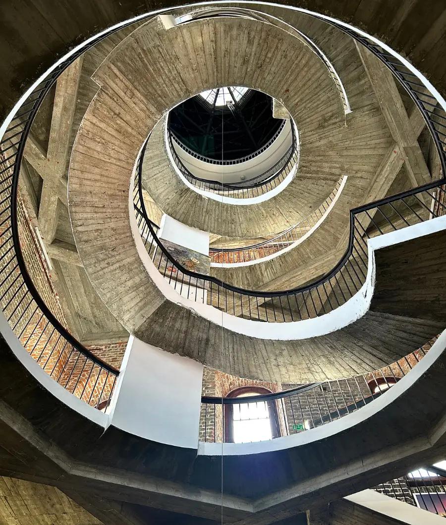Widok w górę monumentalnej spiralnej klatki schodowej z betonu i cegły, ukazujący geometryczne formy i kunszt architektoniczny we wnętrzu zabytkowego budynku.