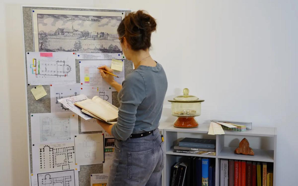 Specjalistka analizuje dokumentację konserwatorską przy tablicy z planami architektonicznymi i notatkami, pracując nad programem prac konserwatorskich.