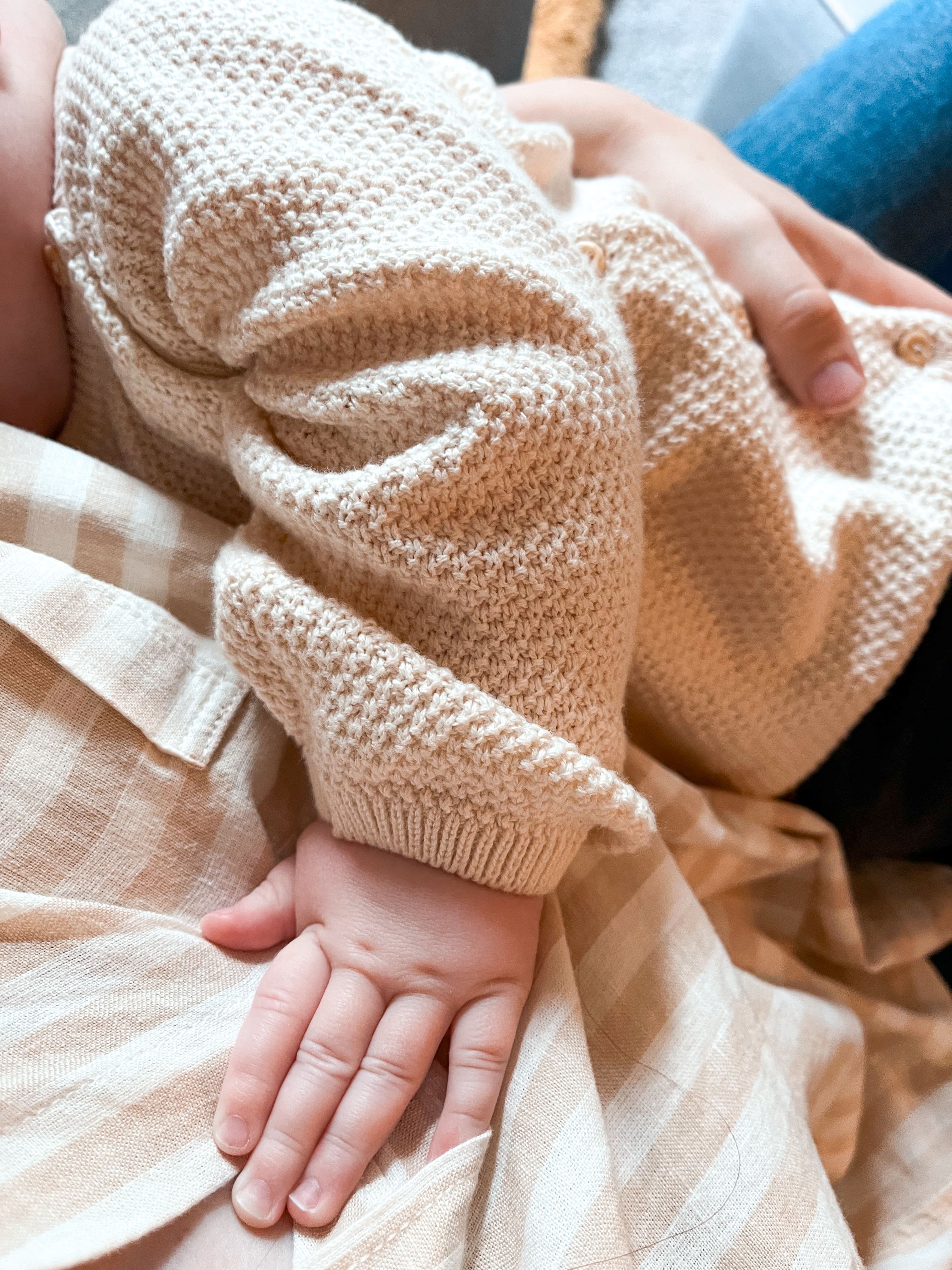 Eine Babyhand auf gestreiftem Stoff neben einem weichen Strickärmel
