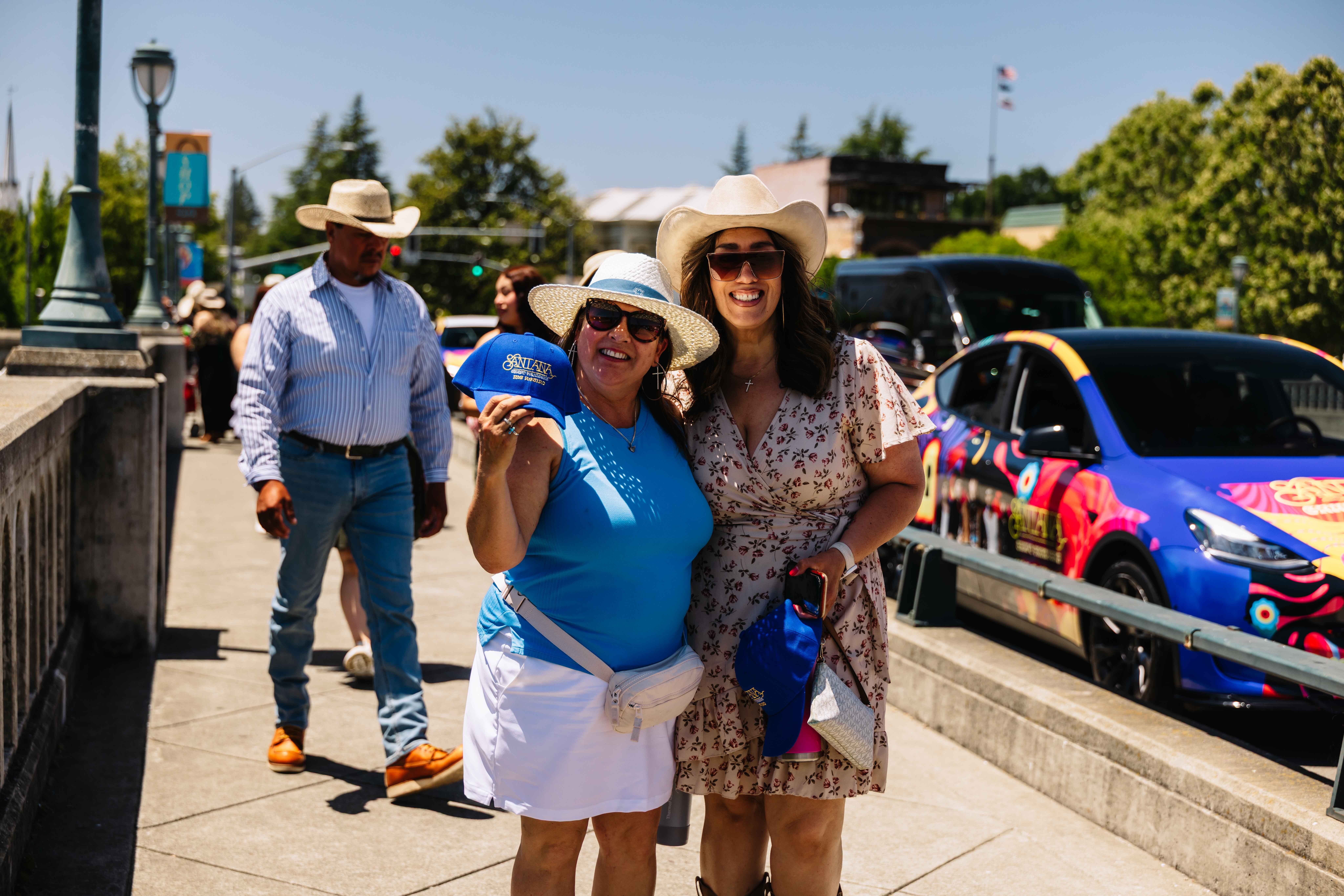 Two smiling women posing with promotional merchandise in front of a colorfully wrapped branded vehicle at an outdoor festival in California