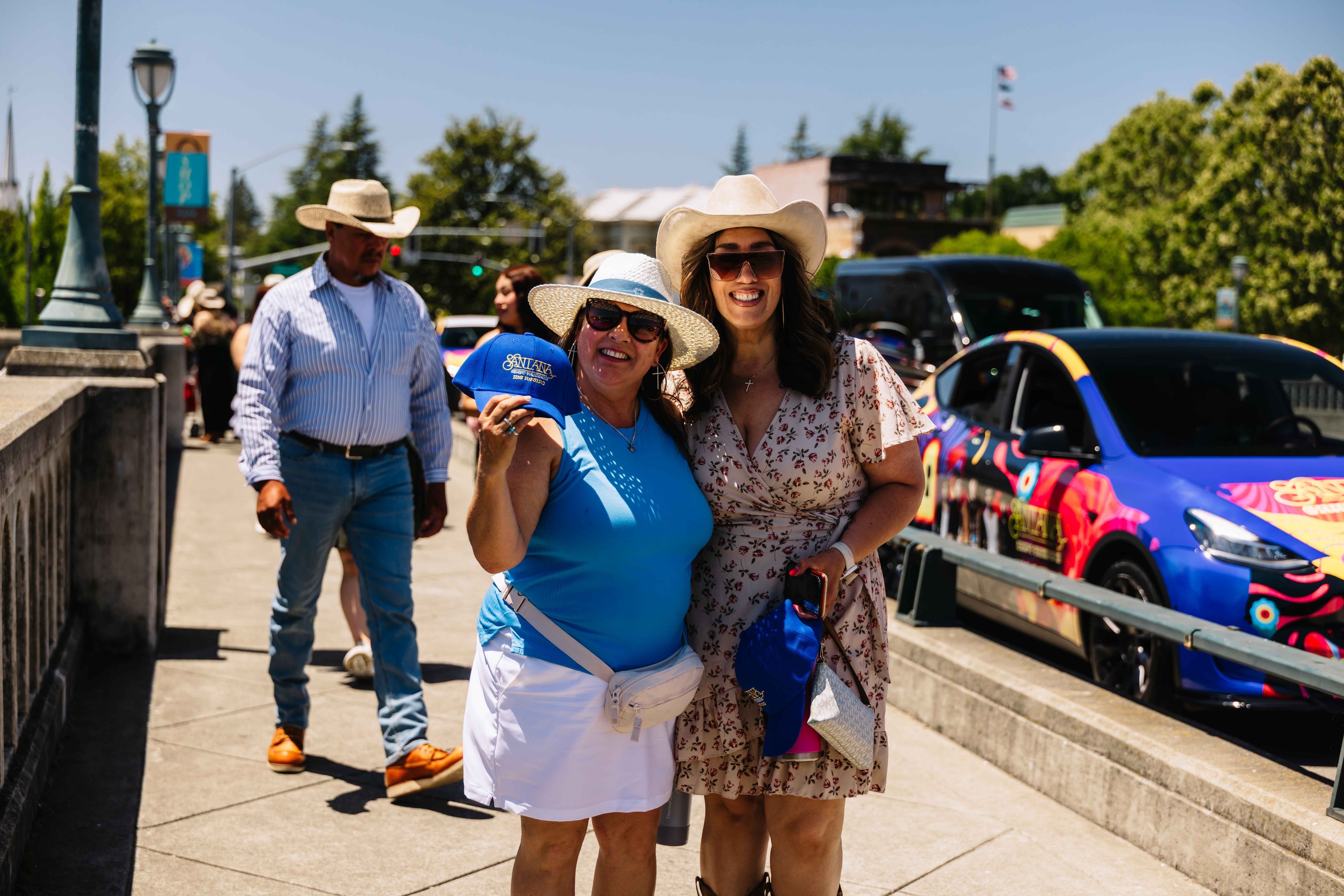 Two smiling women posing with promotional merchandise in front of a colorfully wrapped branded vehicle at an outdoor festival in California